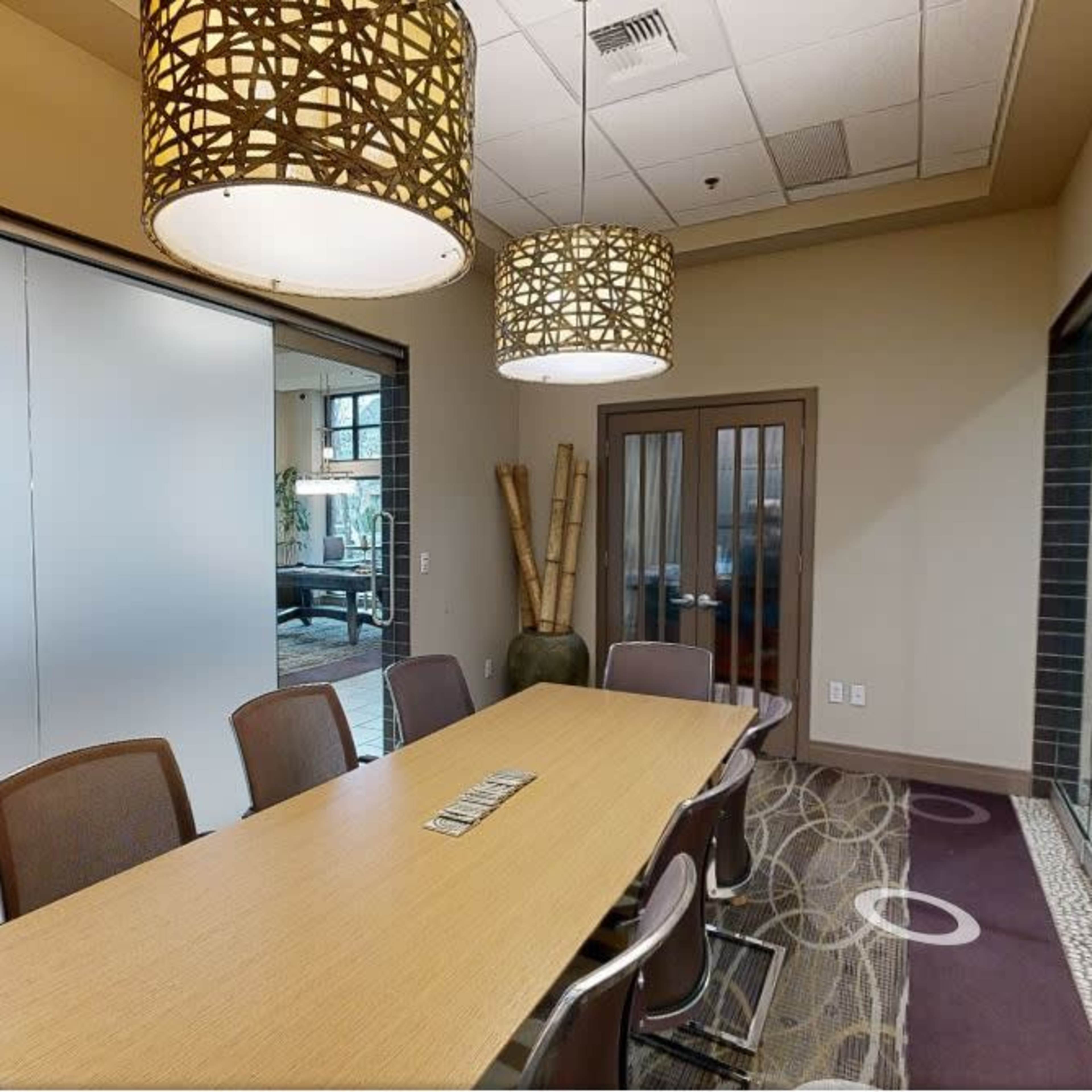 A conference room features a long wooden table surrounded by chairs, with two decorative pendant lights hanging above and glass-paneled doors leading to another space.