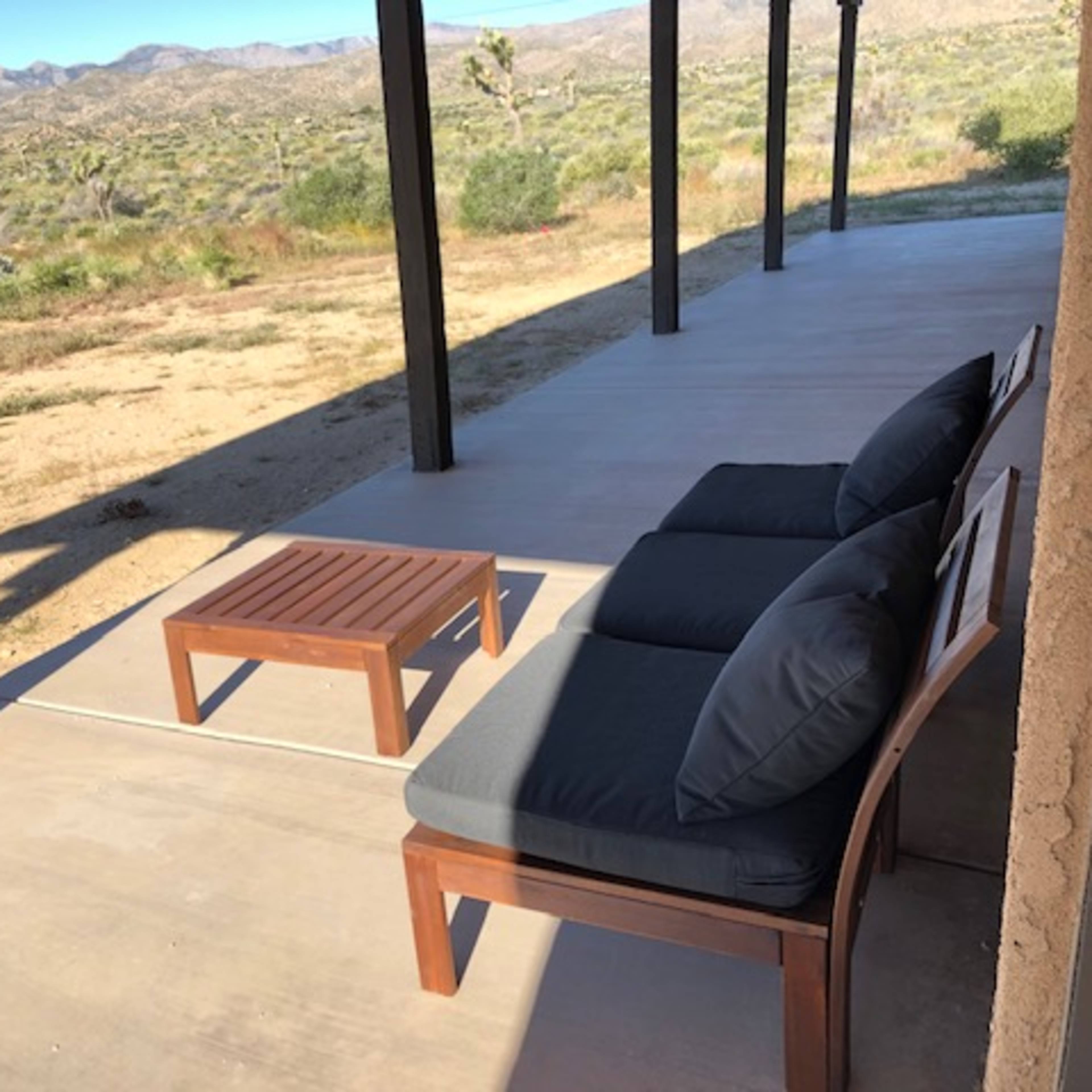 A seating area with a couch and a coffee table is positioned under a porch, overlooking a desert landscape with distant mountains.