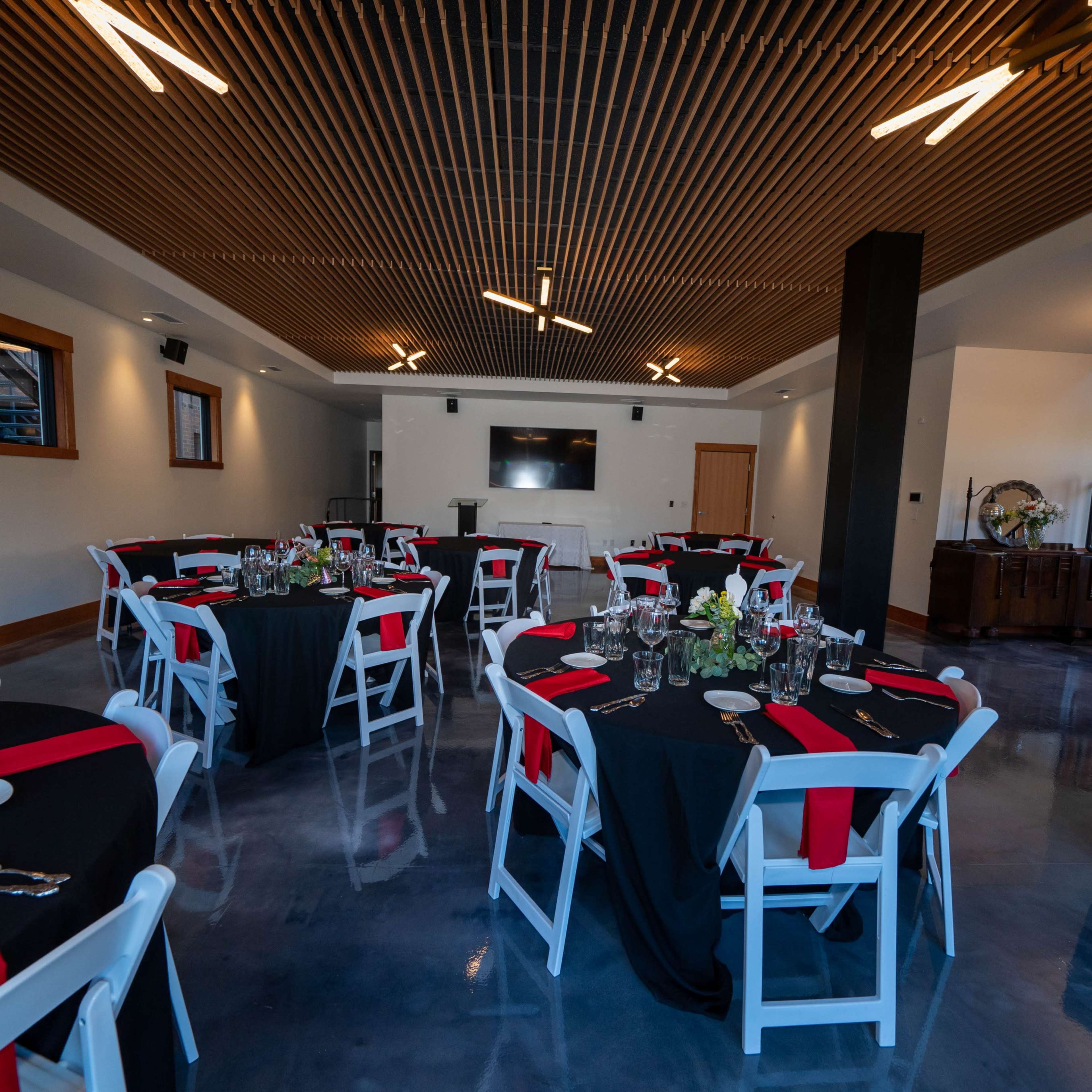 A dining area set with round tables covered in black tablecloths and red accents, arranged around a central space with a flat-screen TV and minimalist decor.