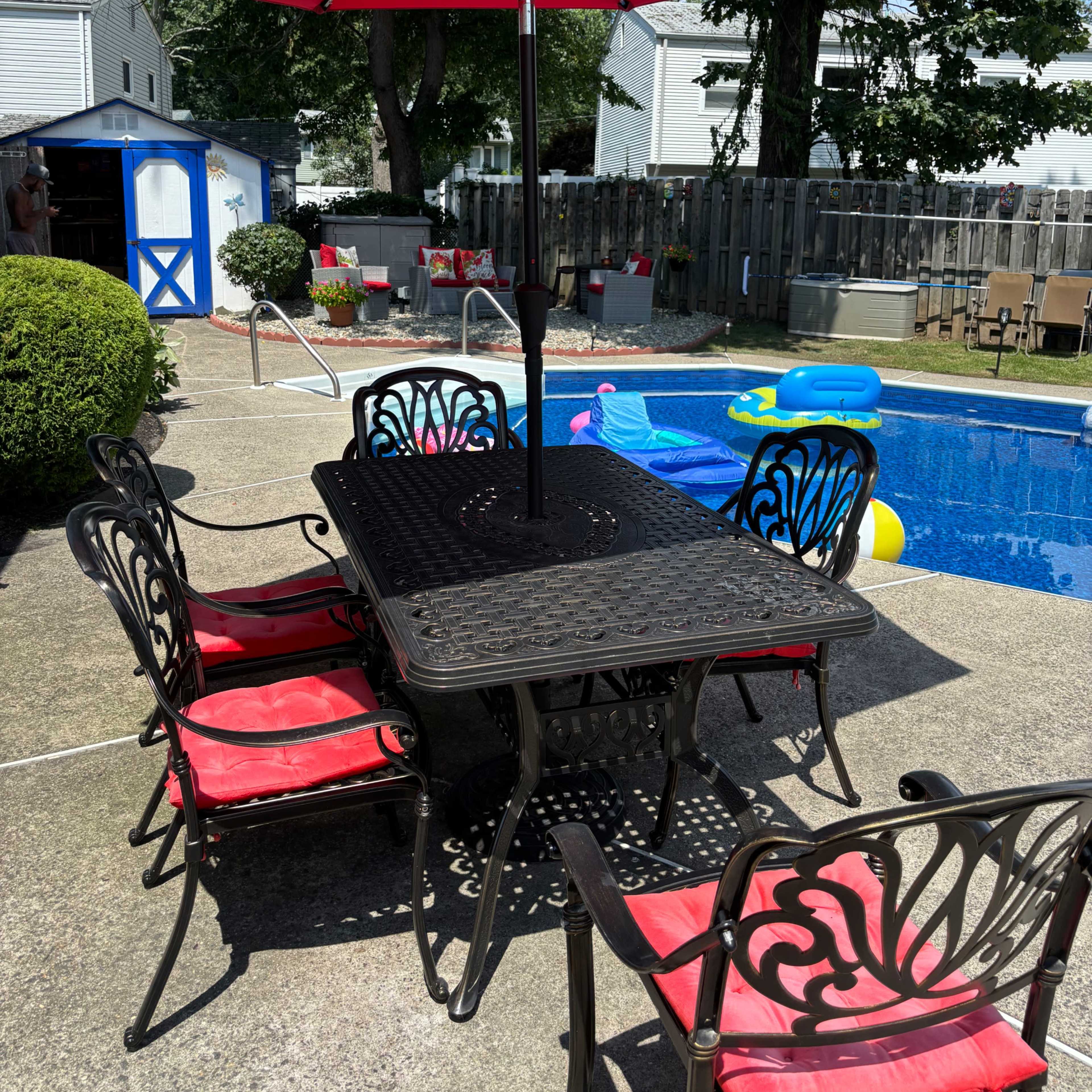 A patio table with red cushions and an umbrella stands next to a swimming pool, surrounded by green bushes and a yard with various items in the background.