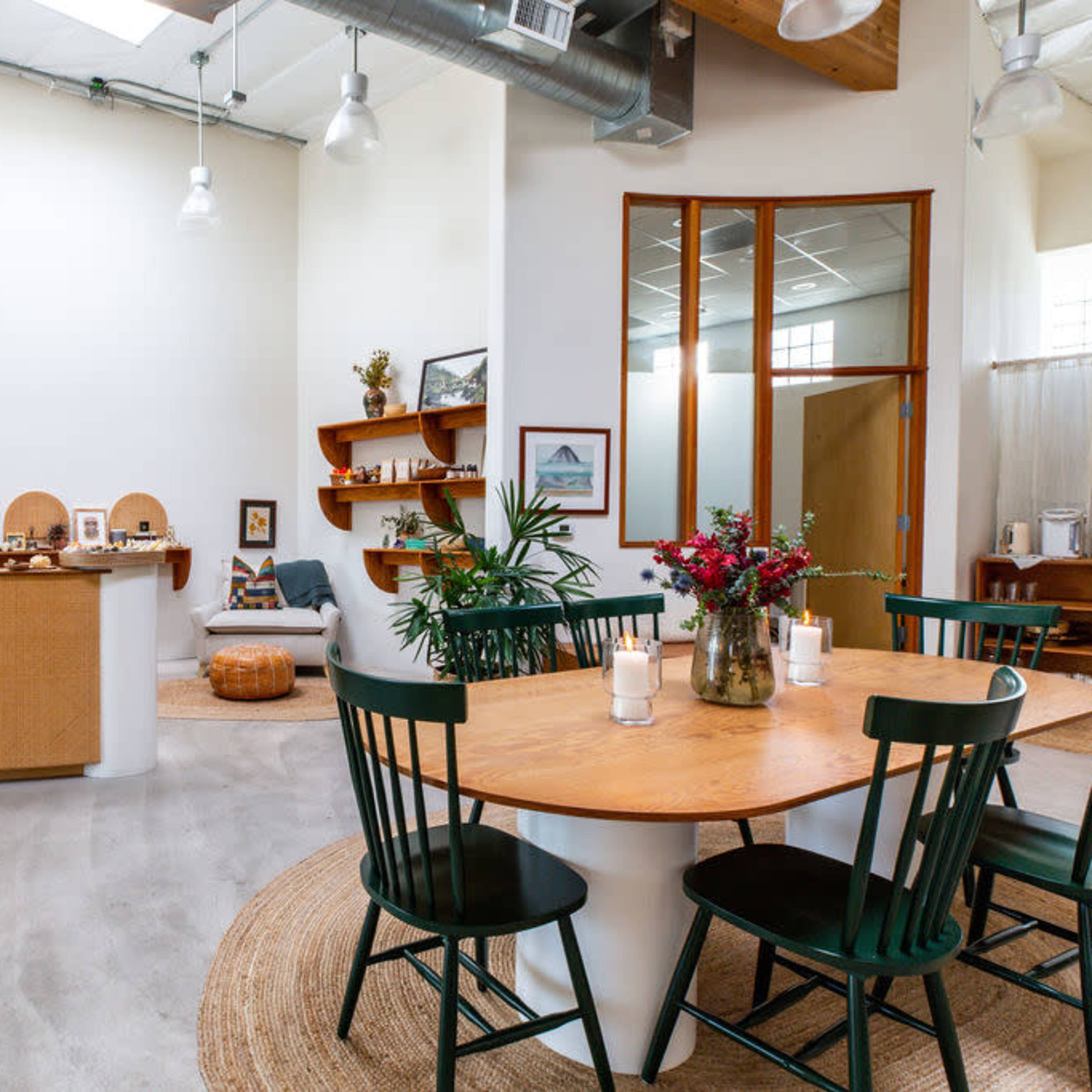 A modern interior of a café or shop featuring a circular wooden table with green chairs, a reception counter, and shelves lined with decorative items.