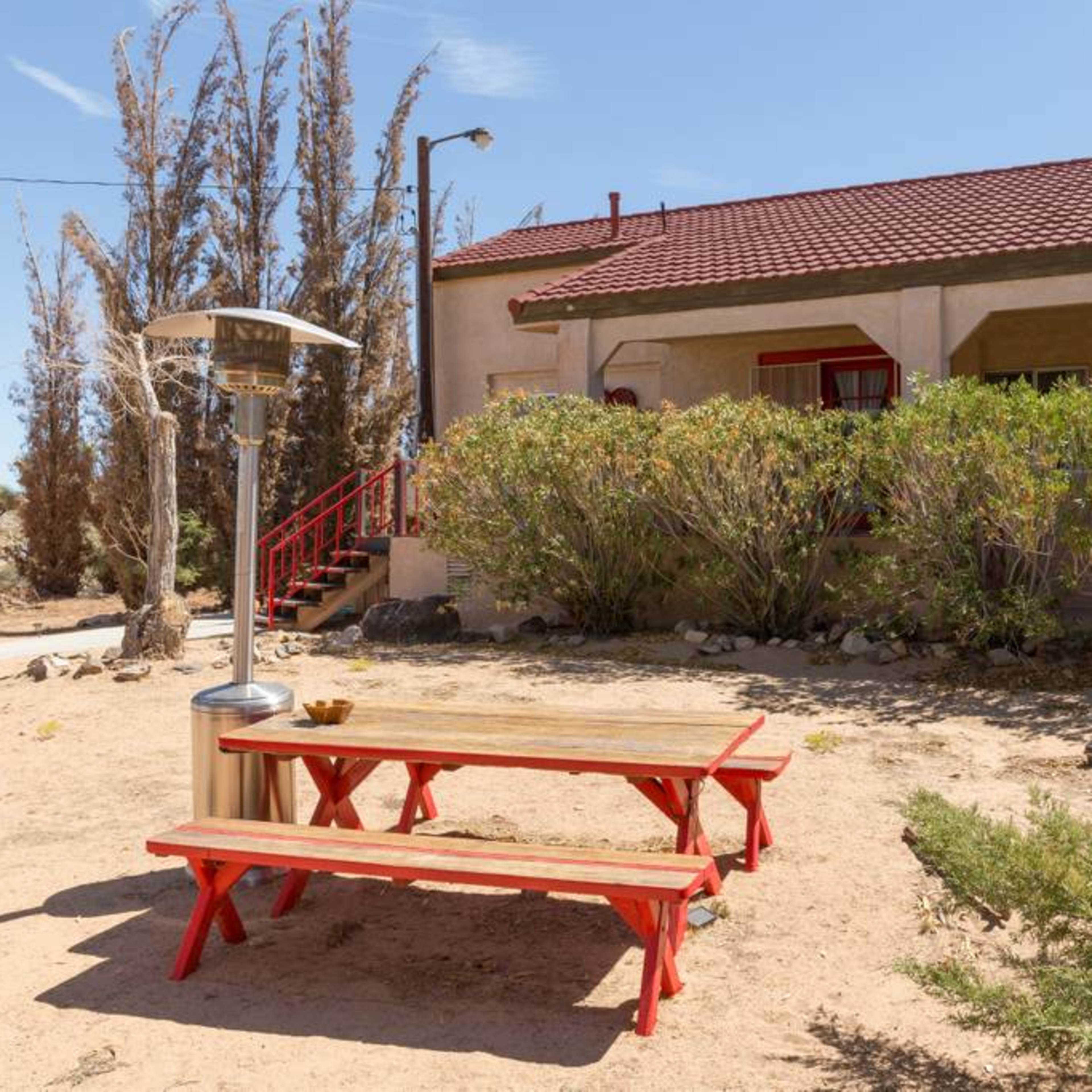 A wooden picnic table with red benches sits in a sandy area next to a building with a red roof and large windows, surrounded by shrubs and barren trees.