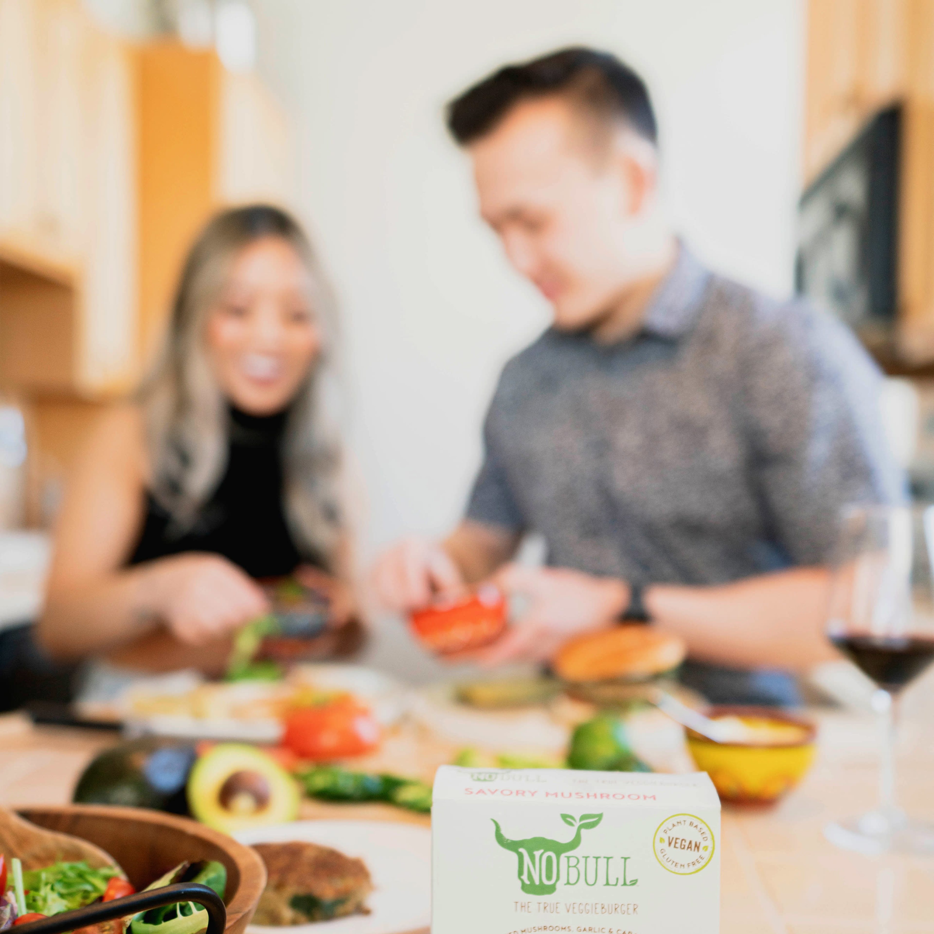 A woman and a man prepare food on a kitchen counter with a box of NoBull Savory Mushroom veggie burgers prominently displayed in the foreground.