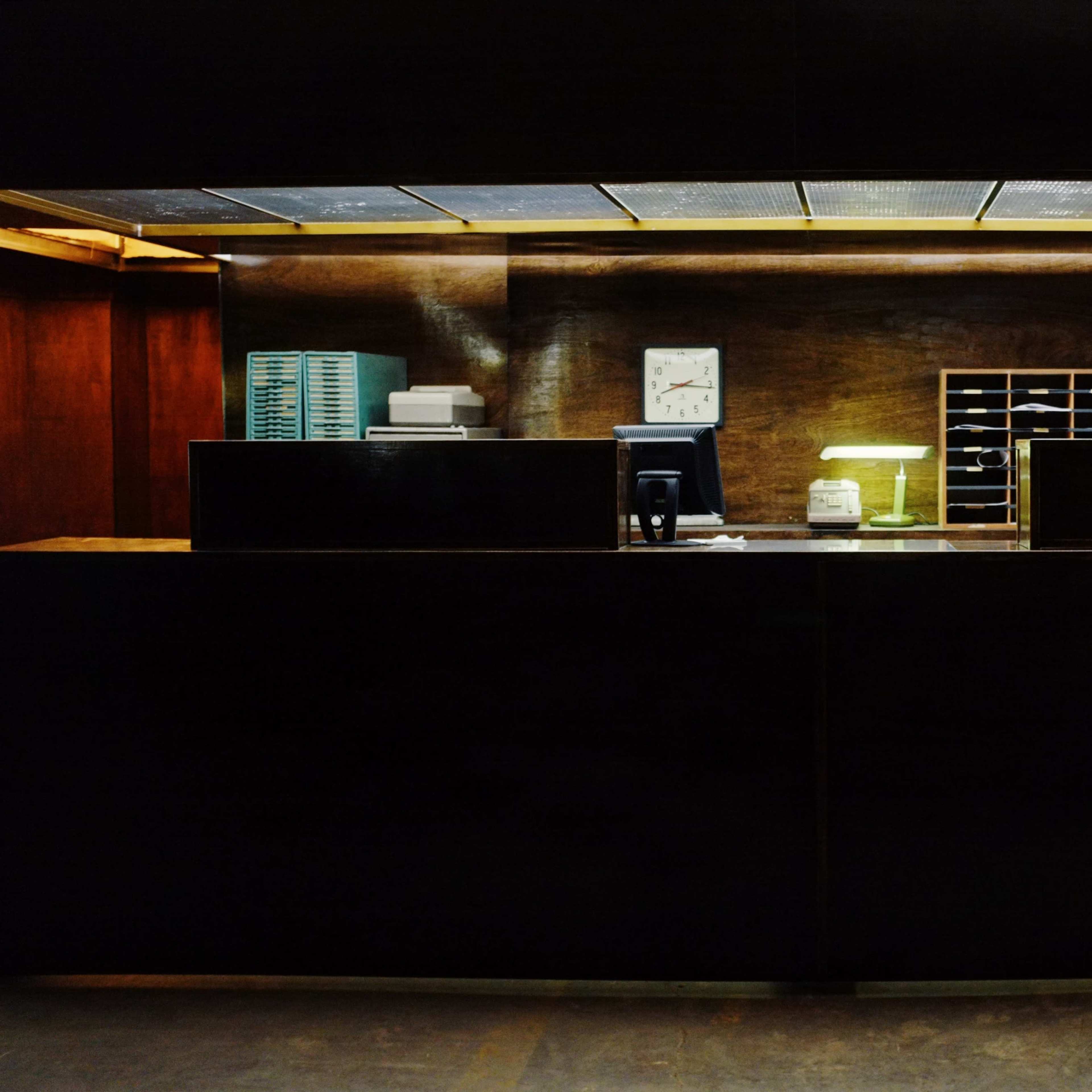 A wooden reception desk with a clock, a lamp, and a stack of items is illuminated against a dark background.