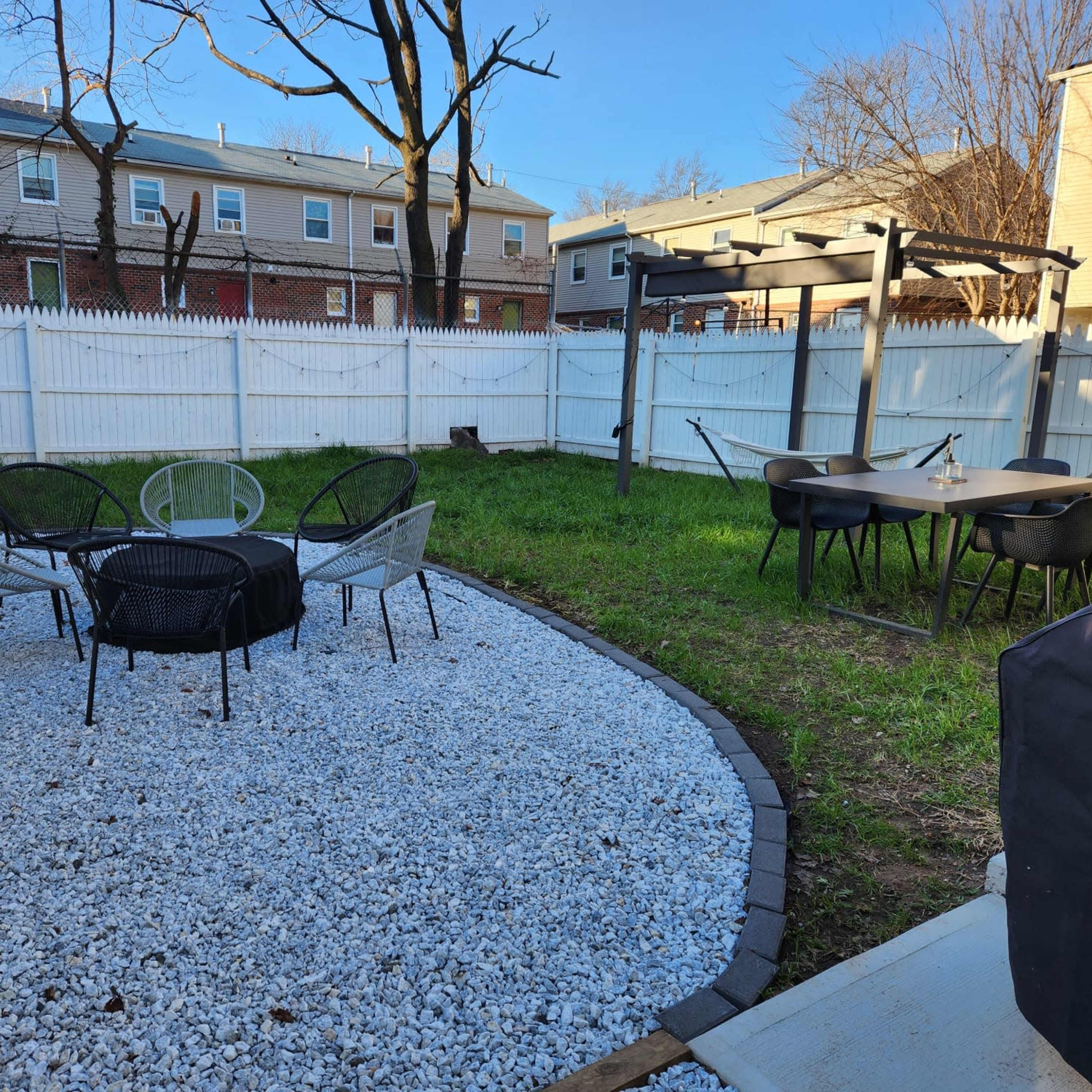 A backyard features a gravel area with a round table and chairs, surrounded by grass and fenced by a white picket fence.