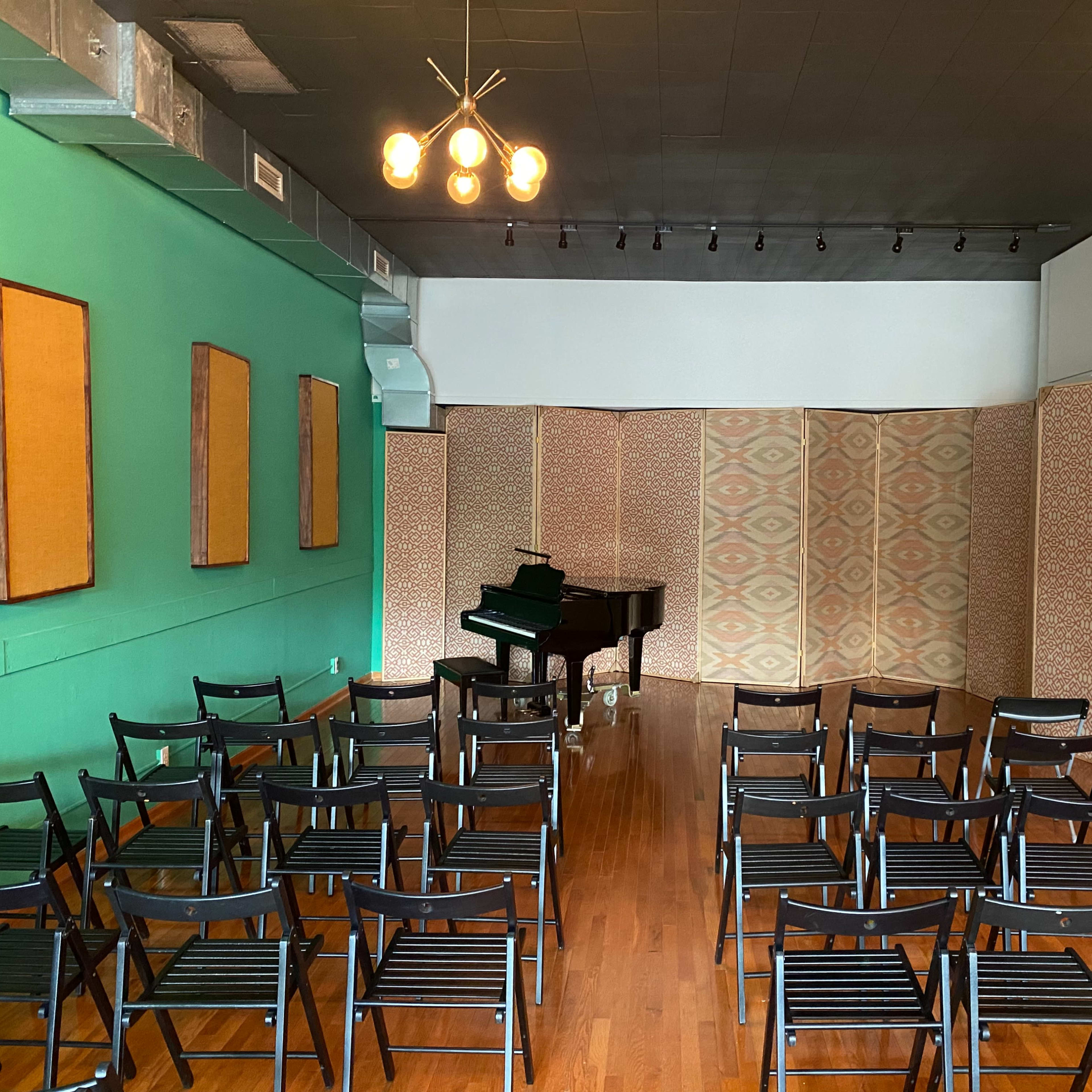 A small performance space with rows of black chairs facing a grand piano against a green wall, flanked by patterned screens.