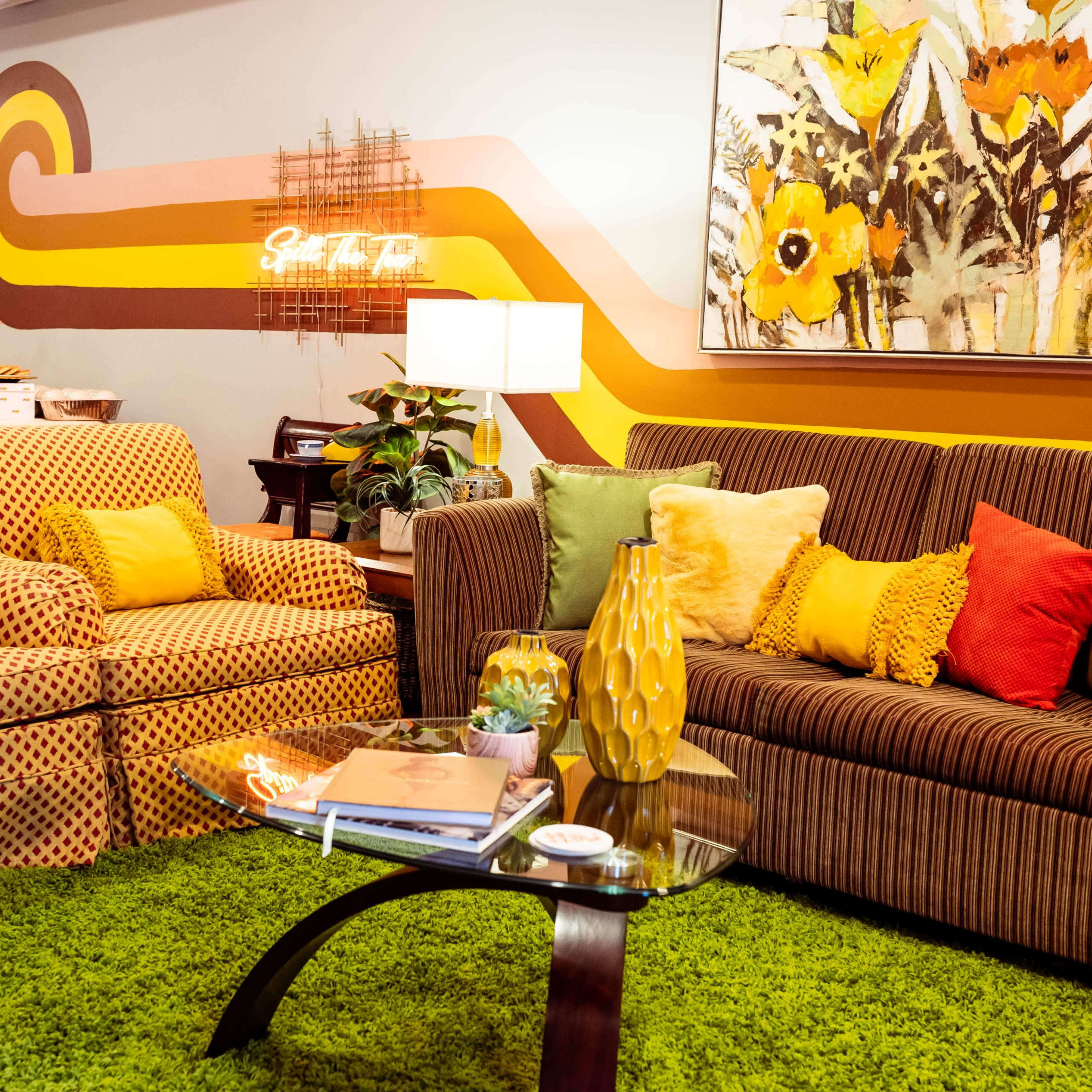 A living room with two sofas, a glass coffee table, and a vibrant, green carpet, set against a decorated wall with colorful patterns and a floral artwork.