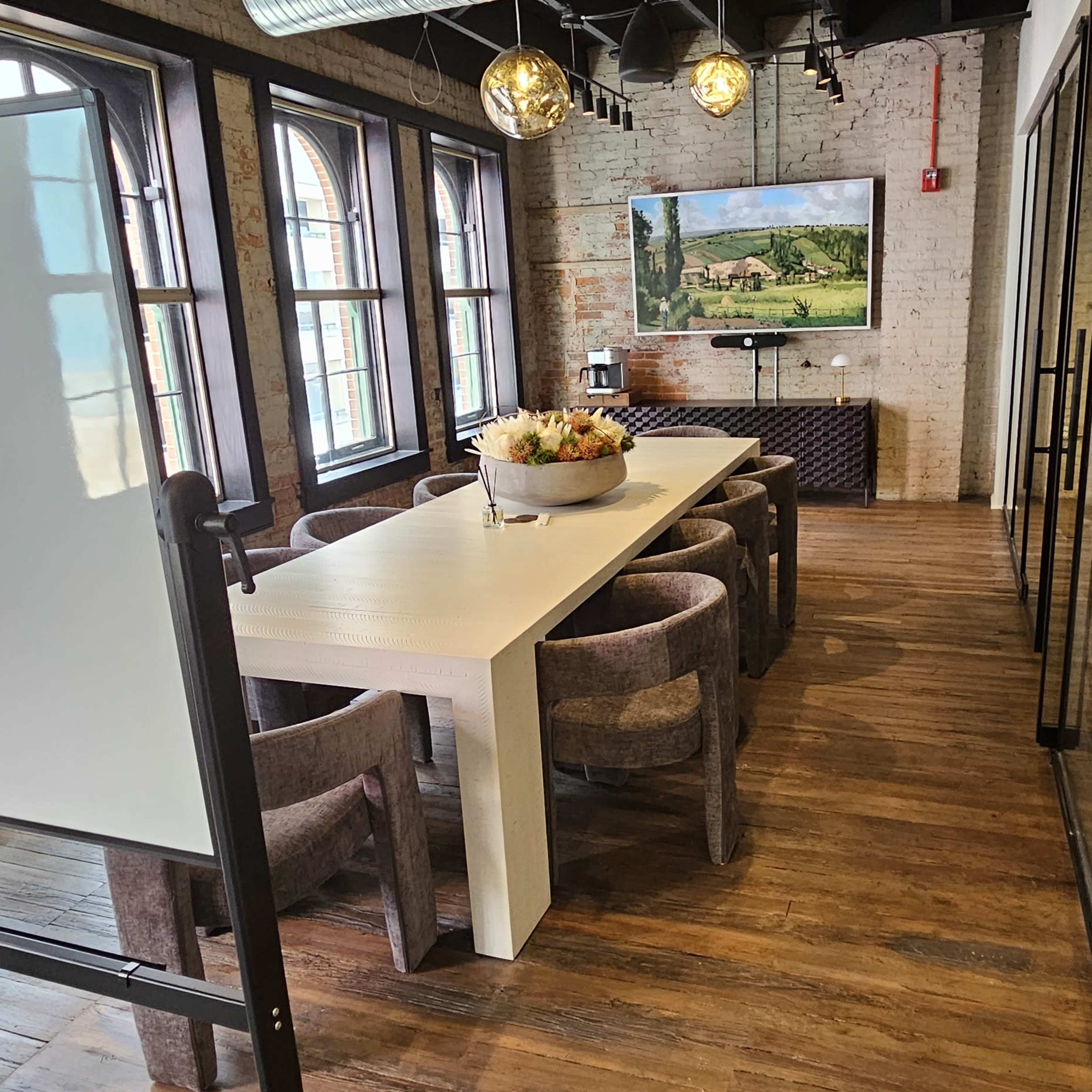 A modern conference room with a white dining table surrounded by brown chairs, a large window, and a television displaying a landscape image.