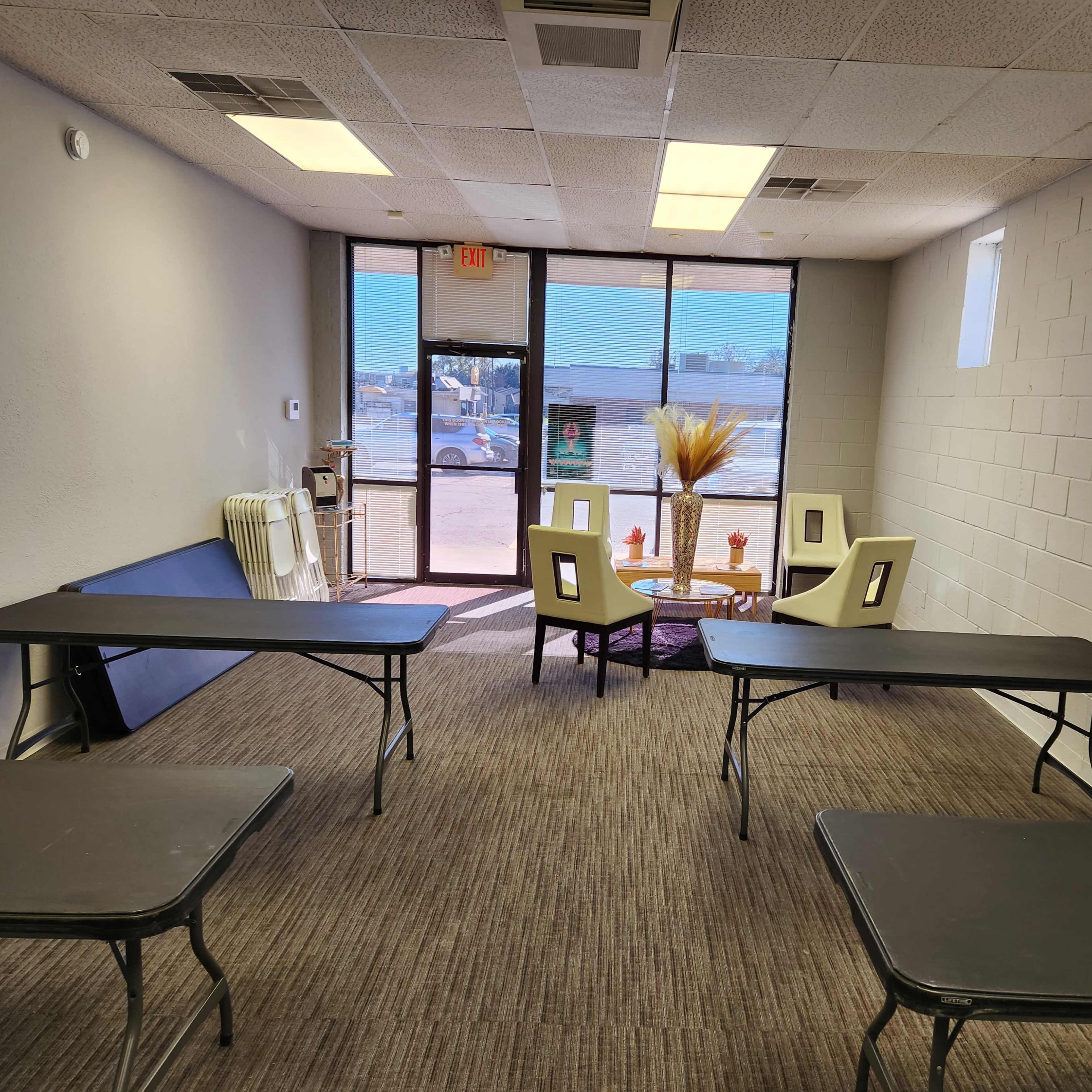 A room with several black tables arranged in a semi-circle, a seating area with chairs and a small table, and a large window letting in natural light.