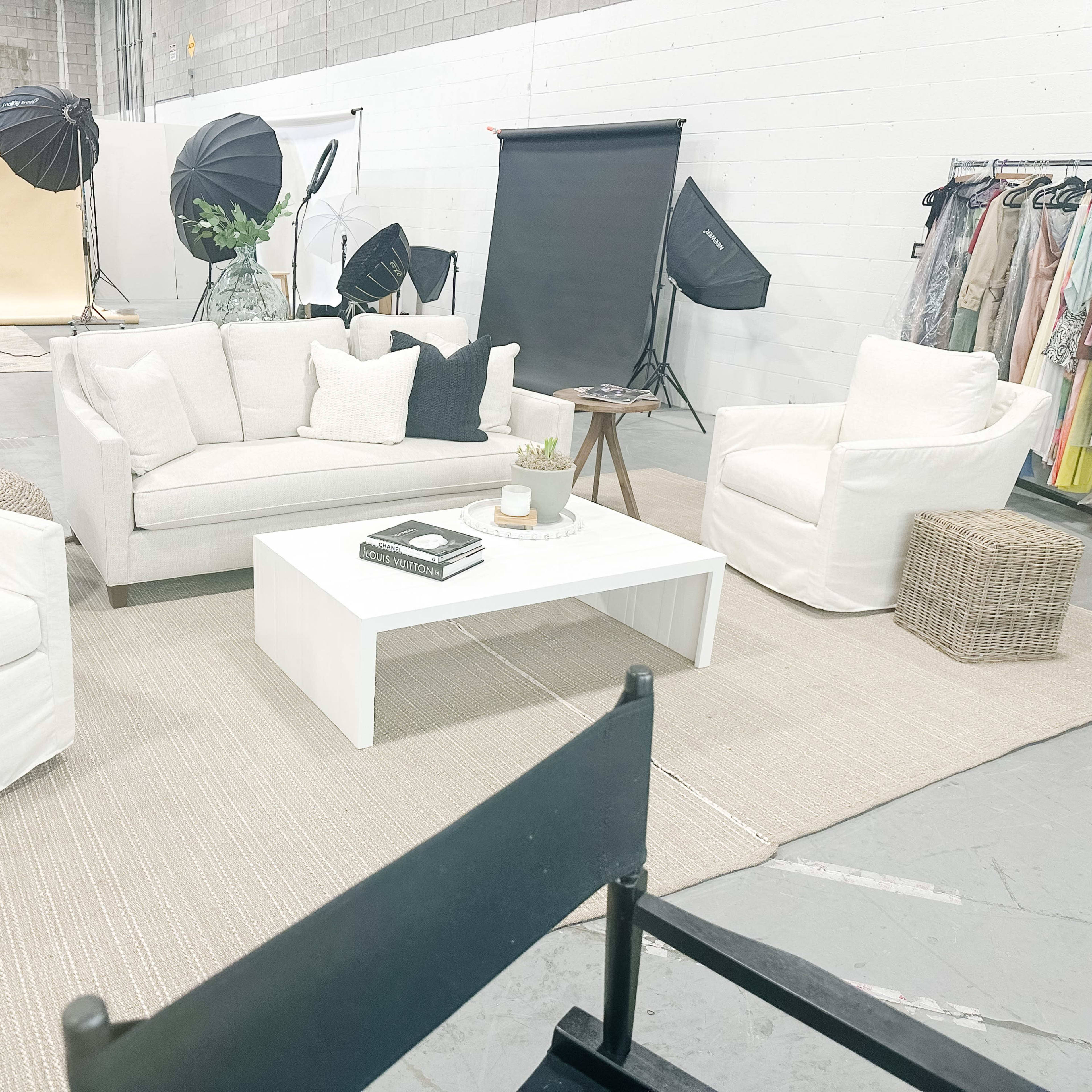 The scene shows a styled photo shoot space with white furniture, a coffee table, and photography equipment set up in a large, industrial-style room.