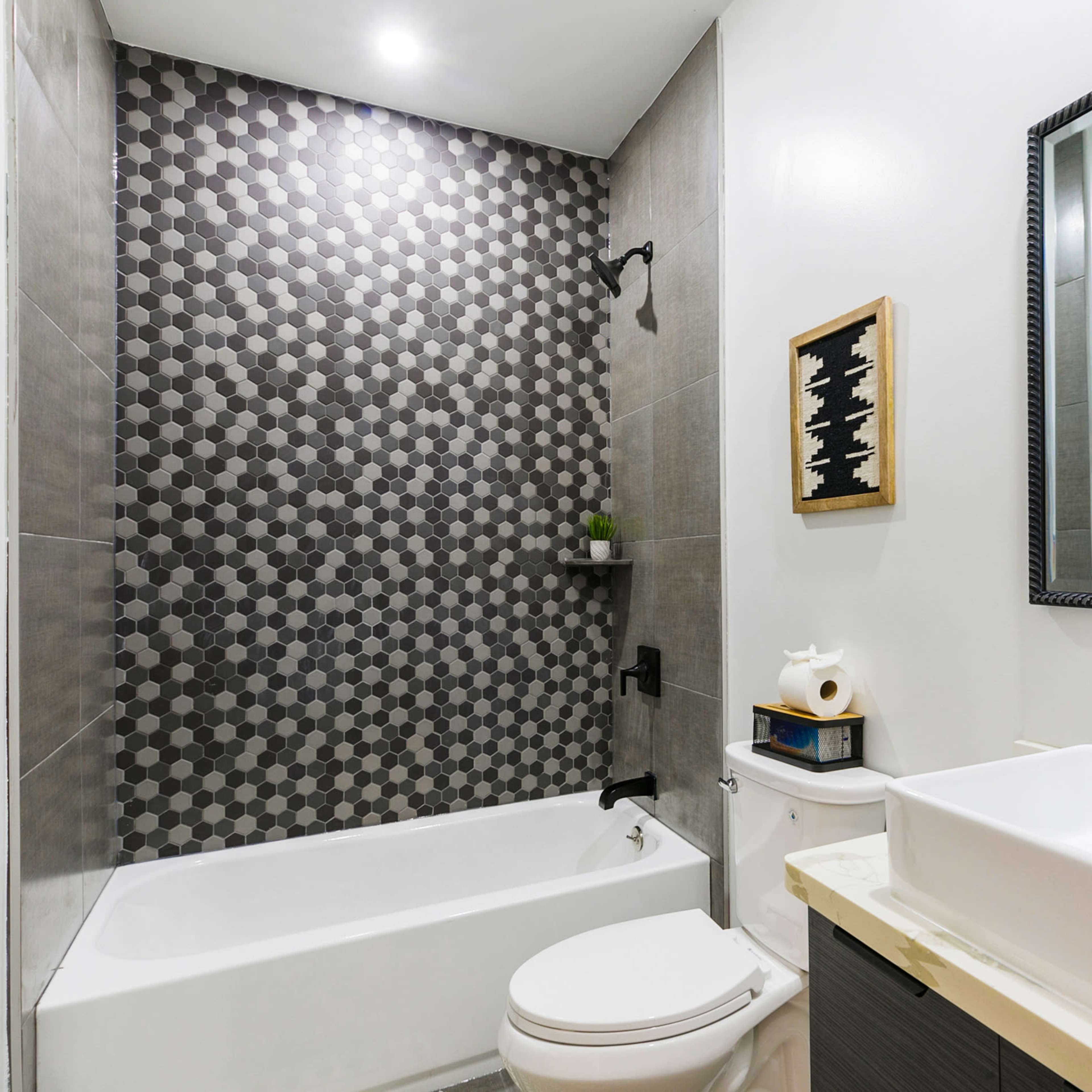 A modern bathroom featuring a bathtub with a hexagonal tiled accent wall, a rectangular sink on a countertop, and a large mirror.