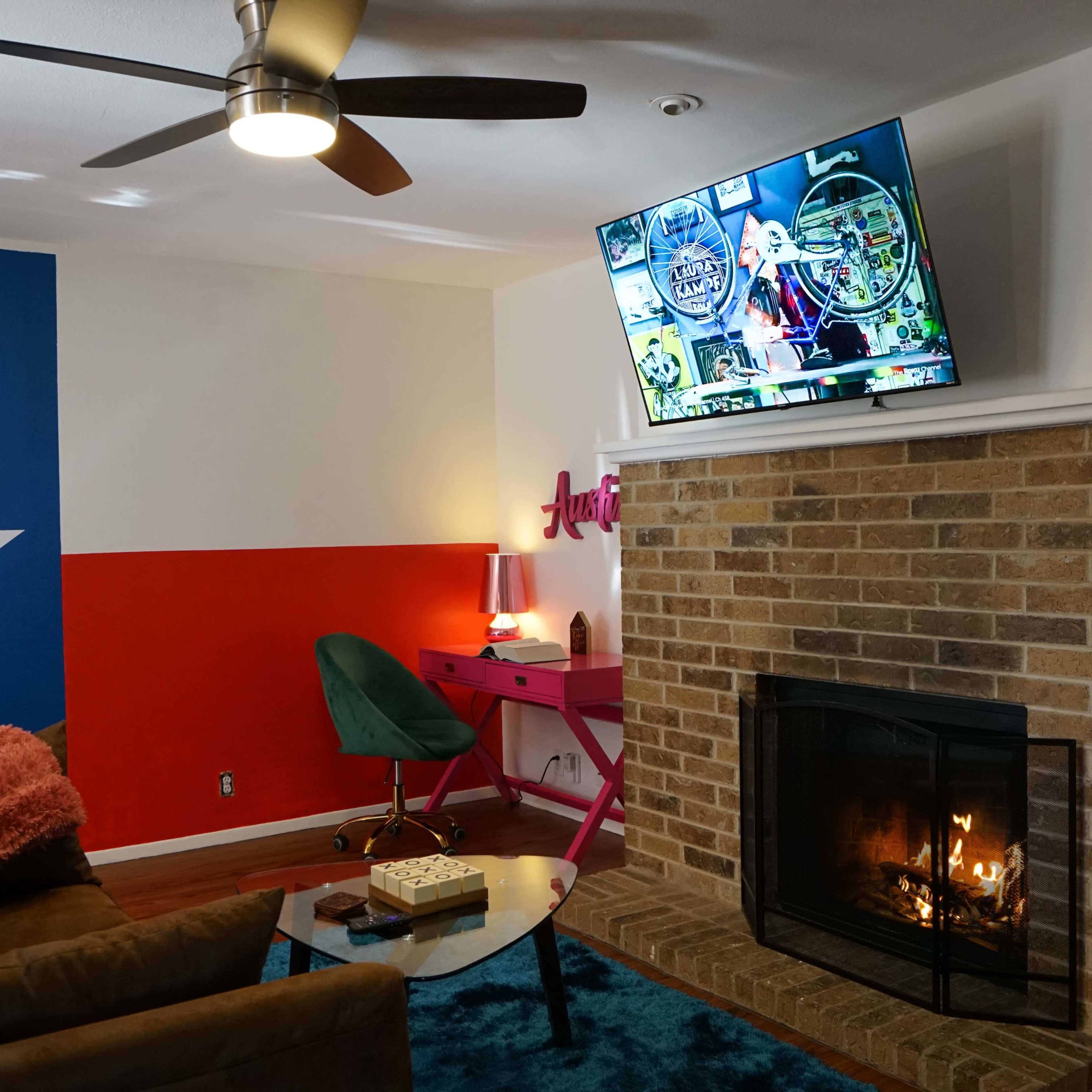 The living room features a Texas flag painted wall, a brick fireplace, a television mounted above it, and a green chair beside a pink desk.