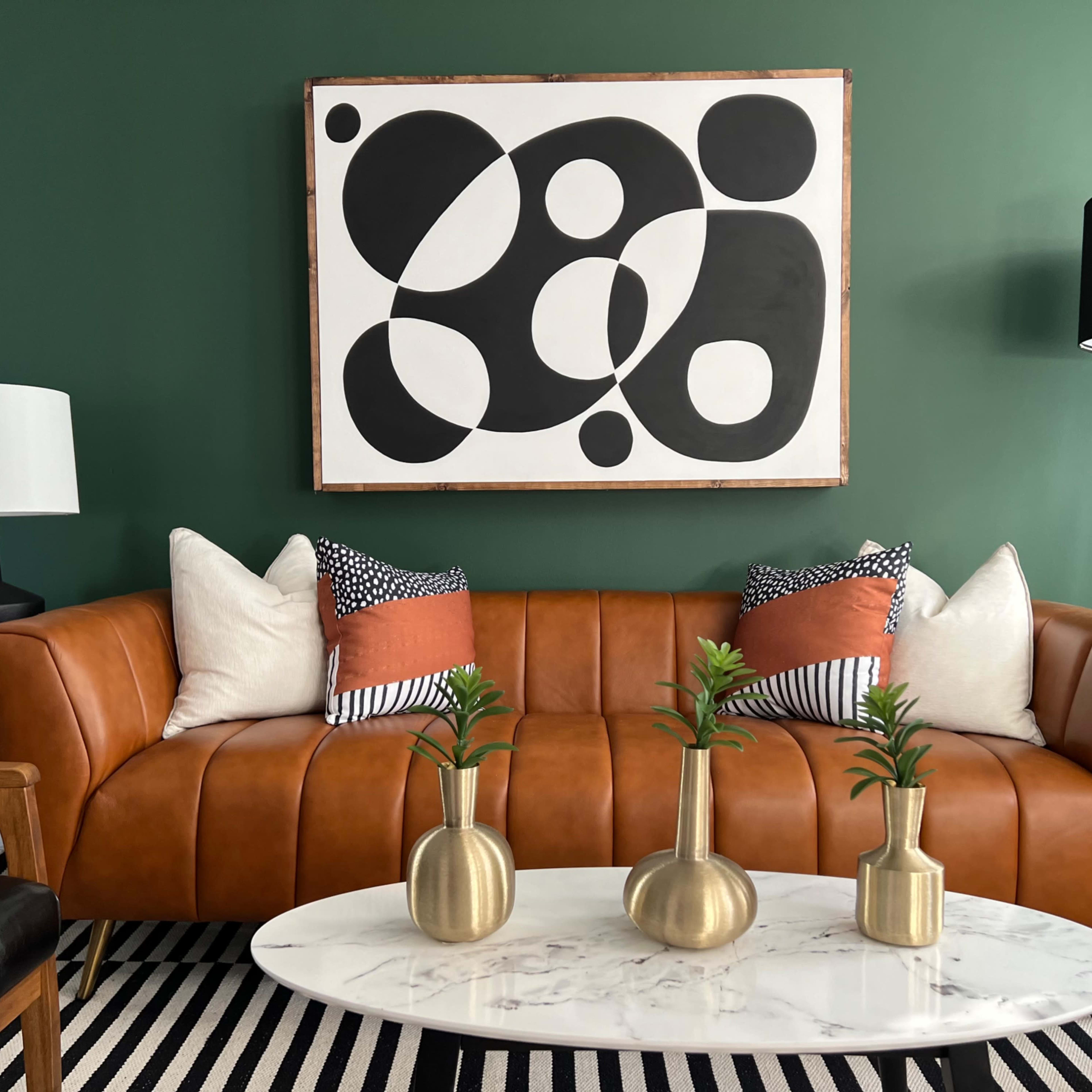 A modern living room with a brown leather sofa, decorative pillows, a round marble coffee table, and abstract artwork on a green wall.