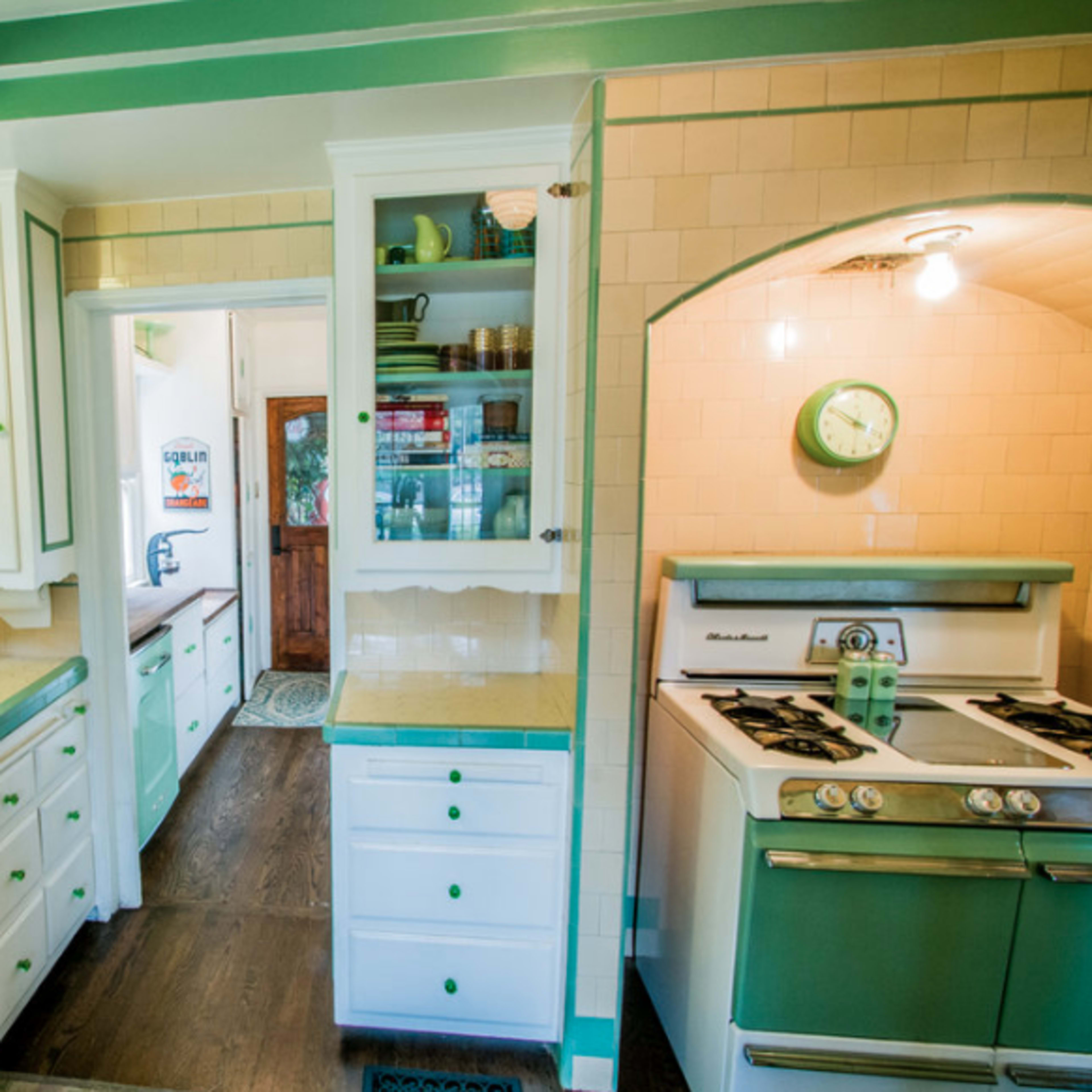 A vintage kitchen with white and green cabinetry, a double sink, and a gas stove, all arranged in a light-filled space with tiled walls.