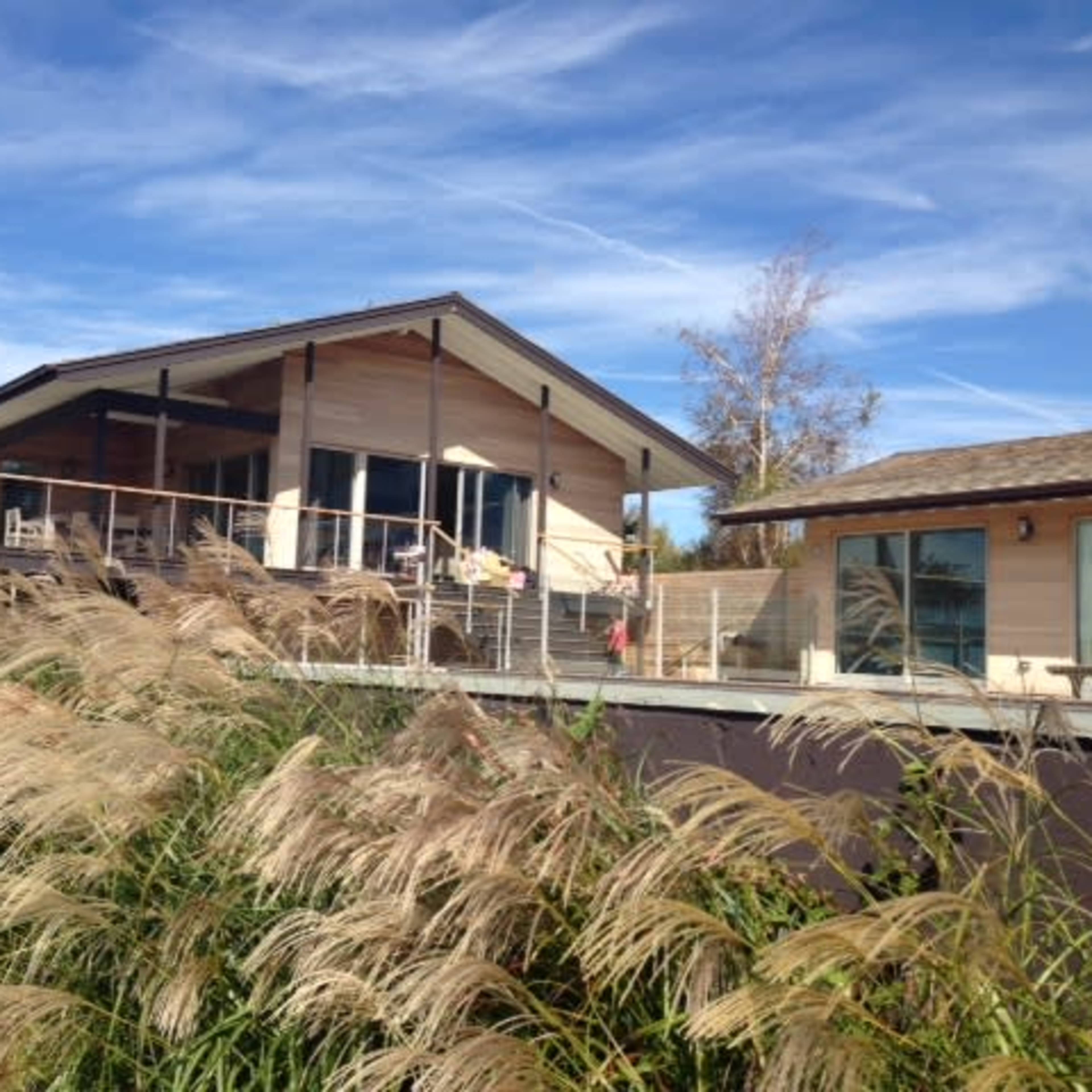 A modern house with a large deck is situated beside tall grass under a clear blue sky.