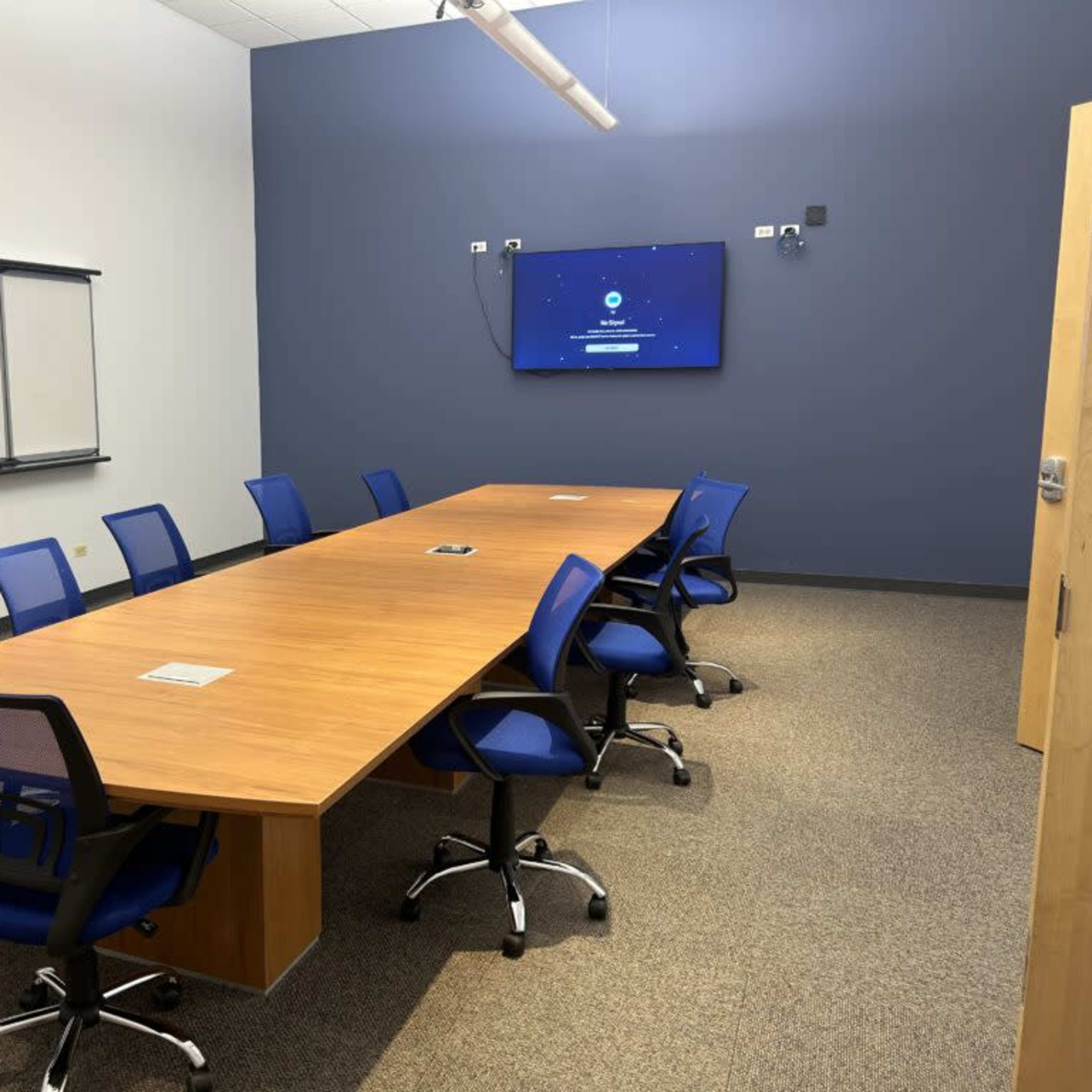 A modern conference room with a long wooden table, blue chairs, and a wall-mounted screen displaying a welcome message.