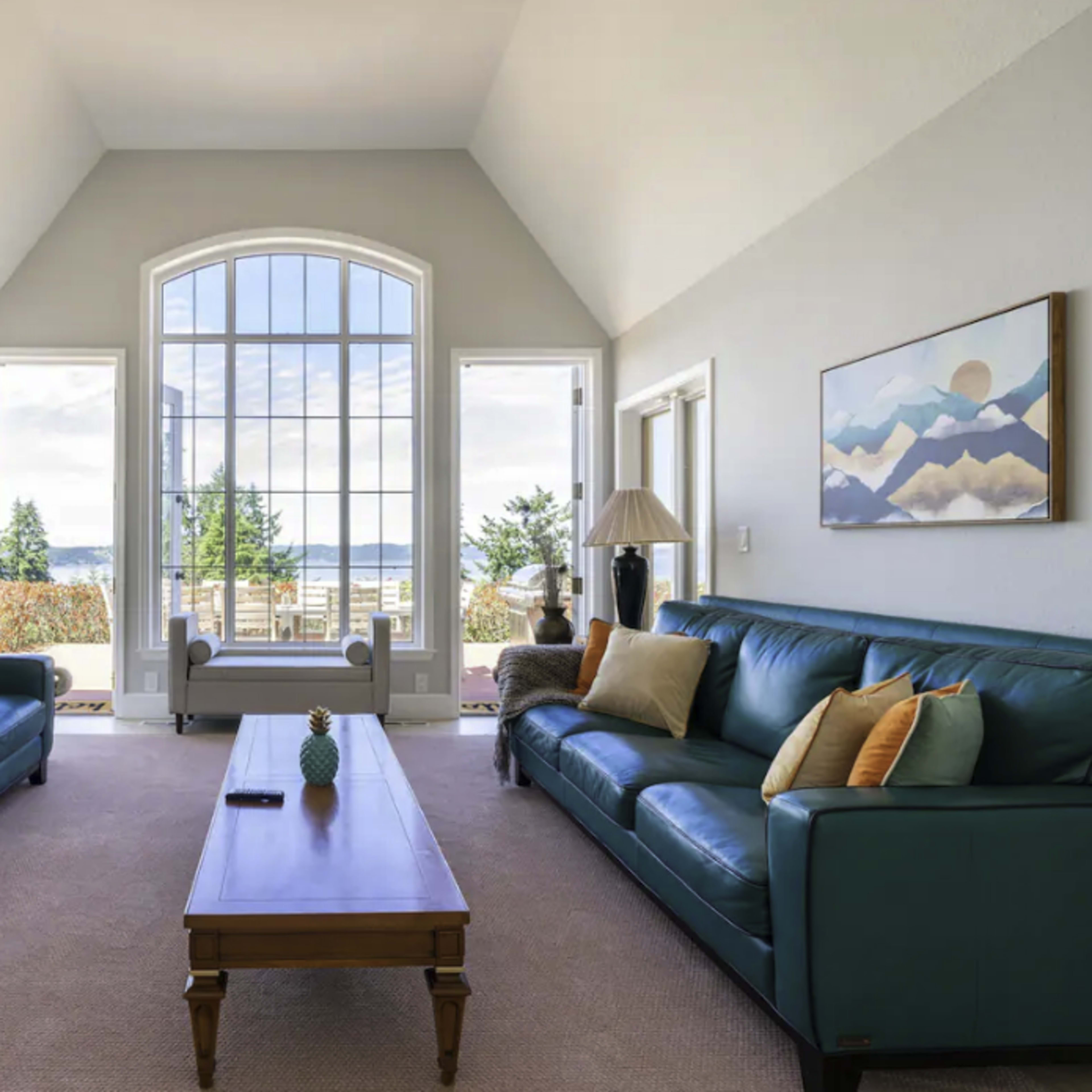 The living room features two teal sofas facing a wooden coffee table, with large windows offering a view of the outdoors.