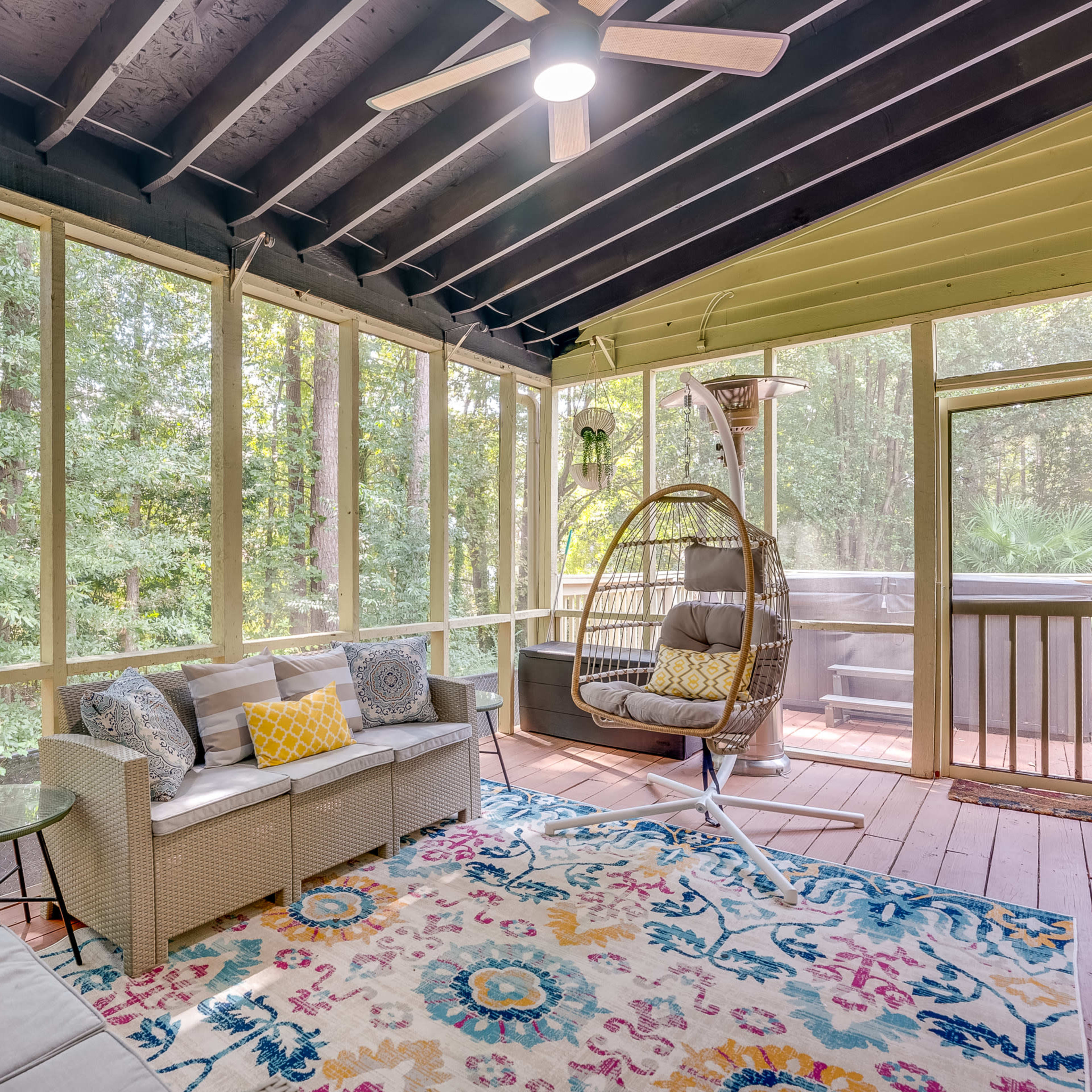 A screened-in porch with a sofa, a hanging chair, and a table, surrounded by trees and decorated with a colorful rug.