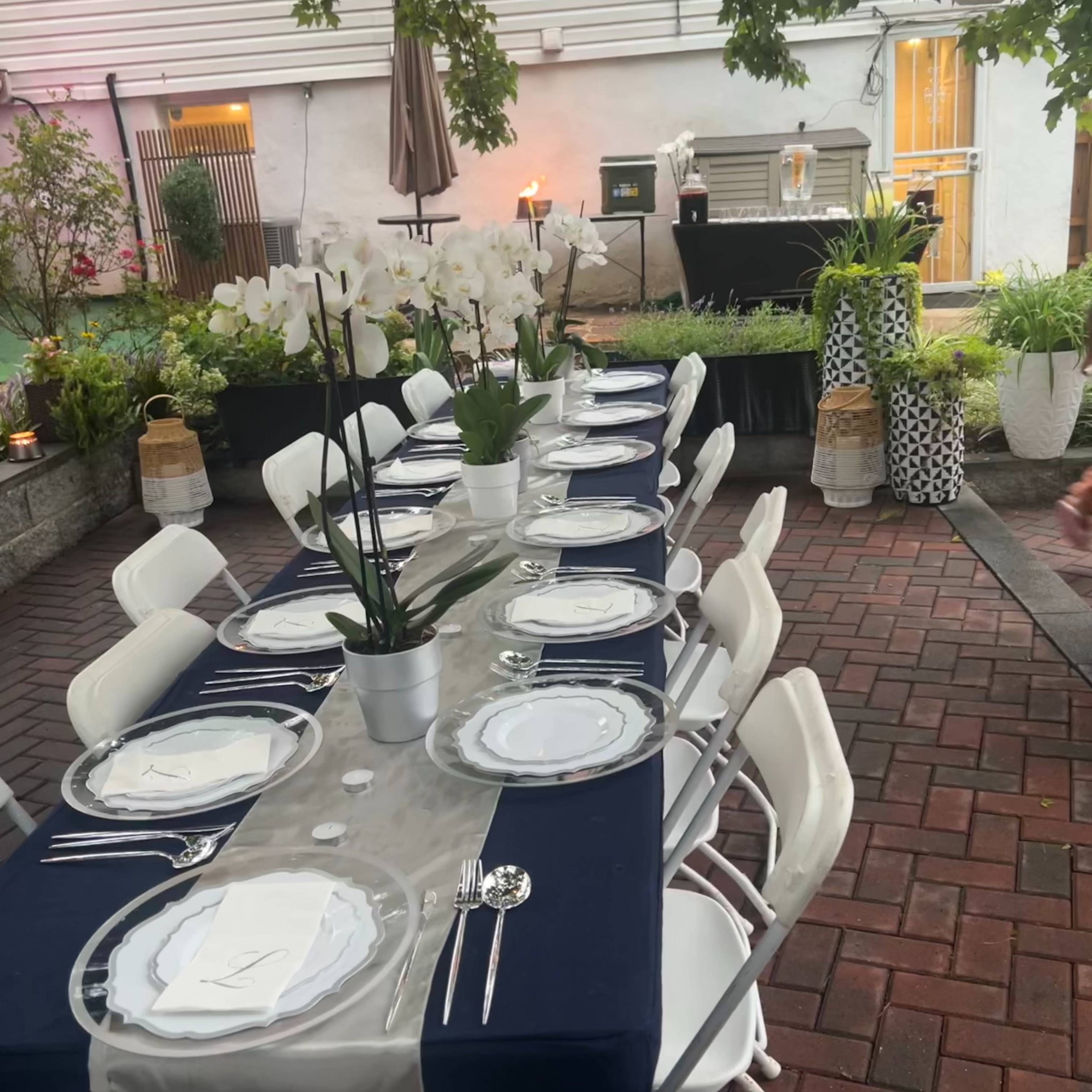 A long outdoor dining table is set with white plates and silverware, surrounded by chairs, under a canopy of greenery and decorative lanterns.