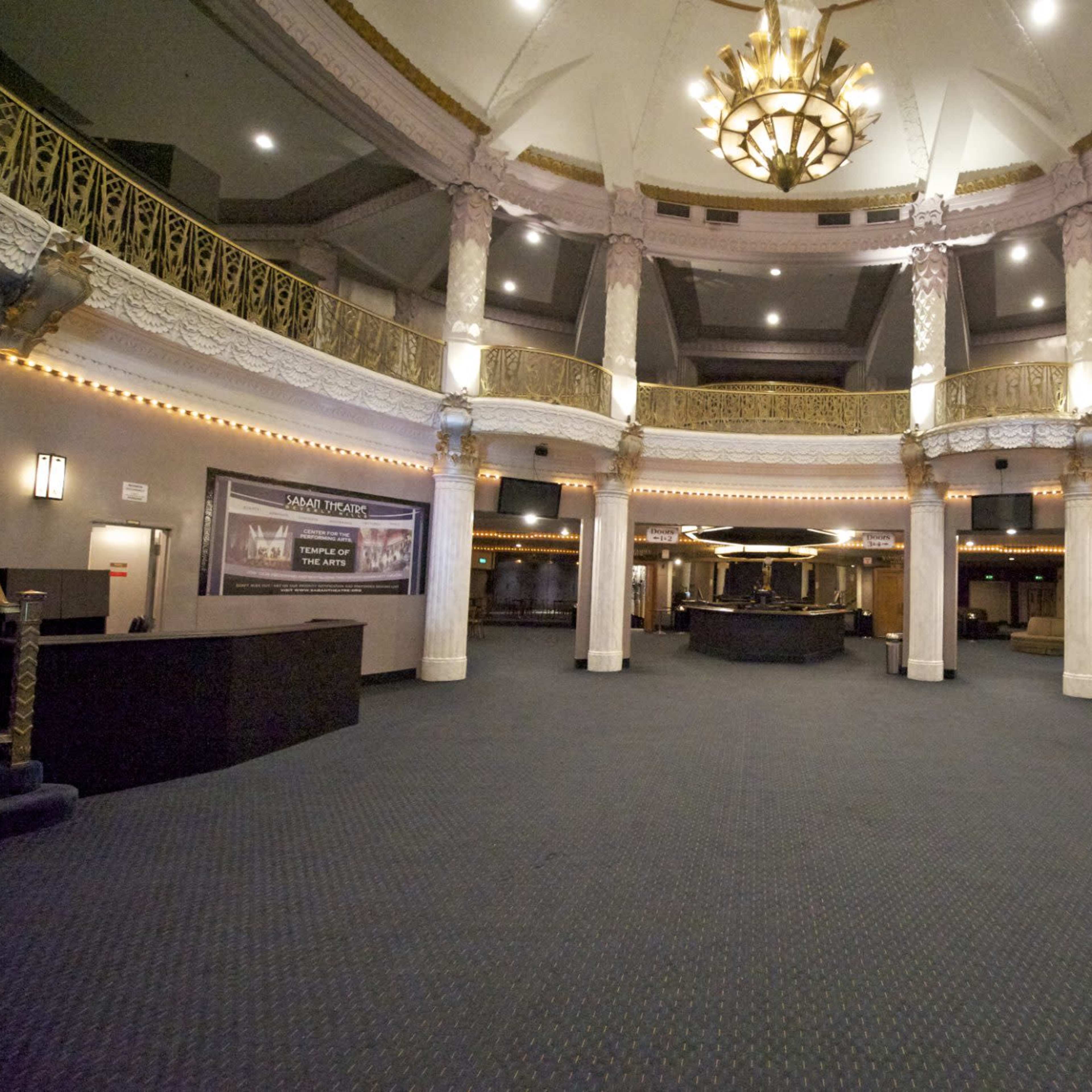 The interior of a theater features grand architectural details, including ornate columns and a large chandelier, with a spacious, empty lobby area.