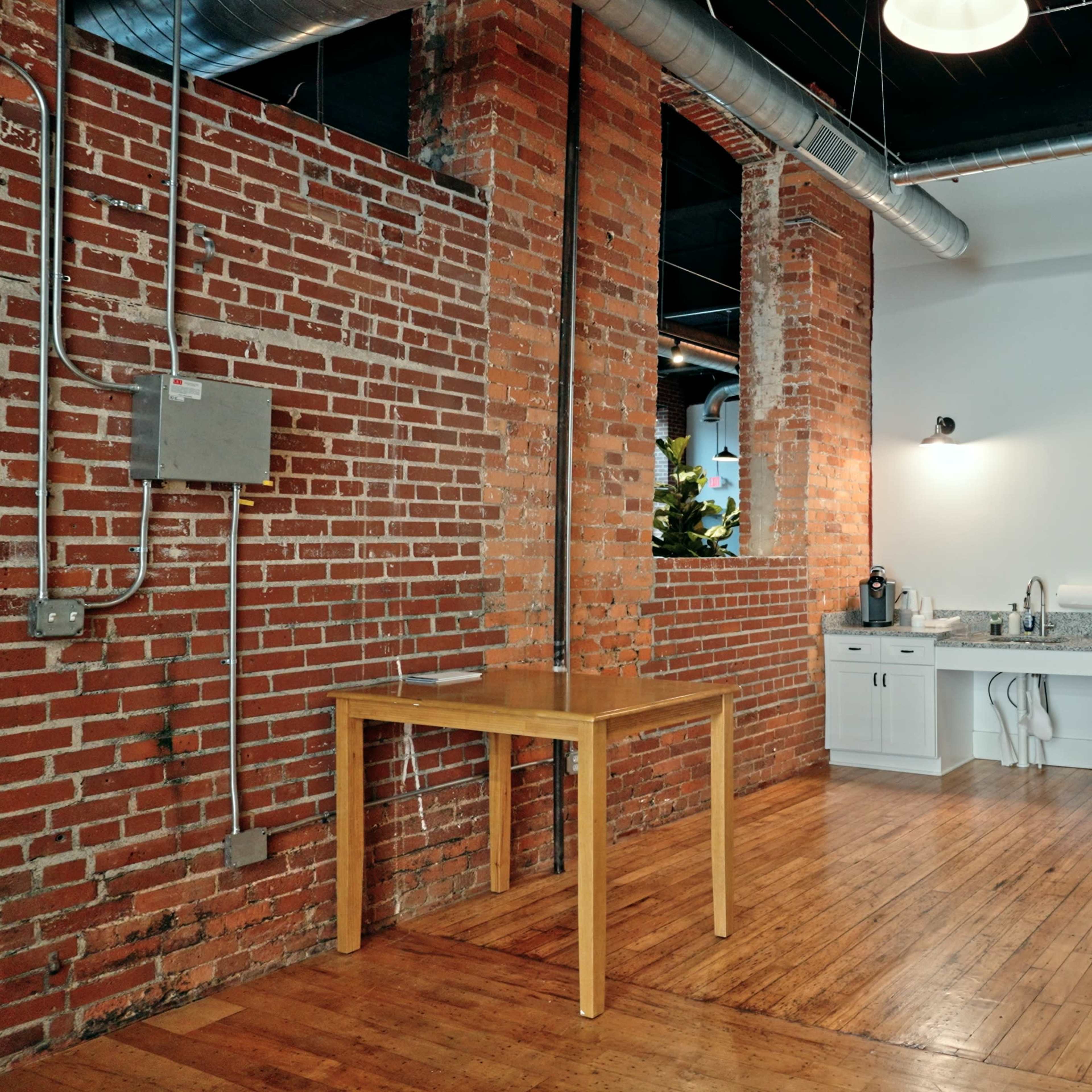 An industrial-style kitchen area featuring exposed brick walls, a wooden table, a countertop with a sink, and various appliances including a refrigerator and microwave.