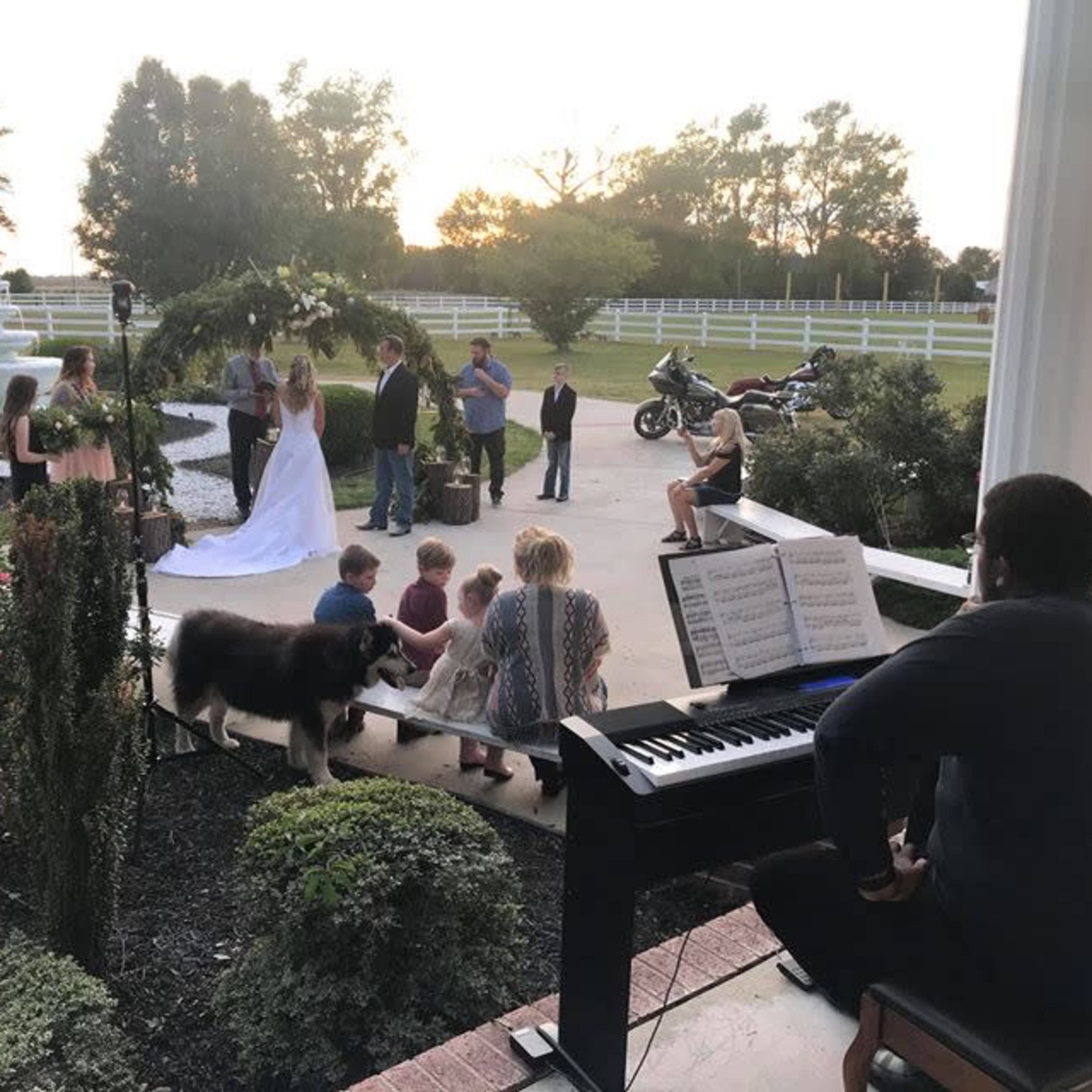 A wedding ceremony takes place outdoors near a fountain, with guests gathered and a musician playing piano.