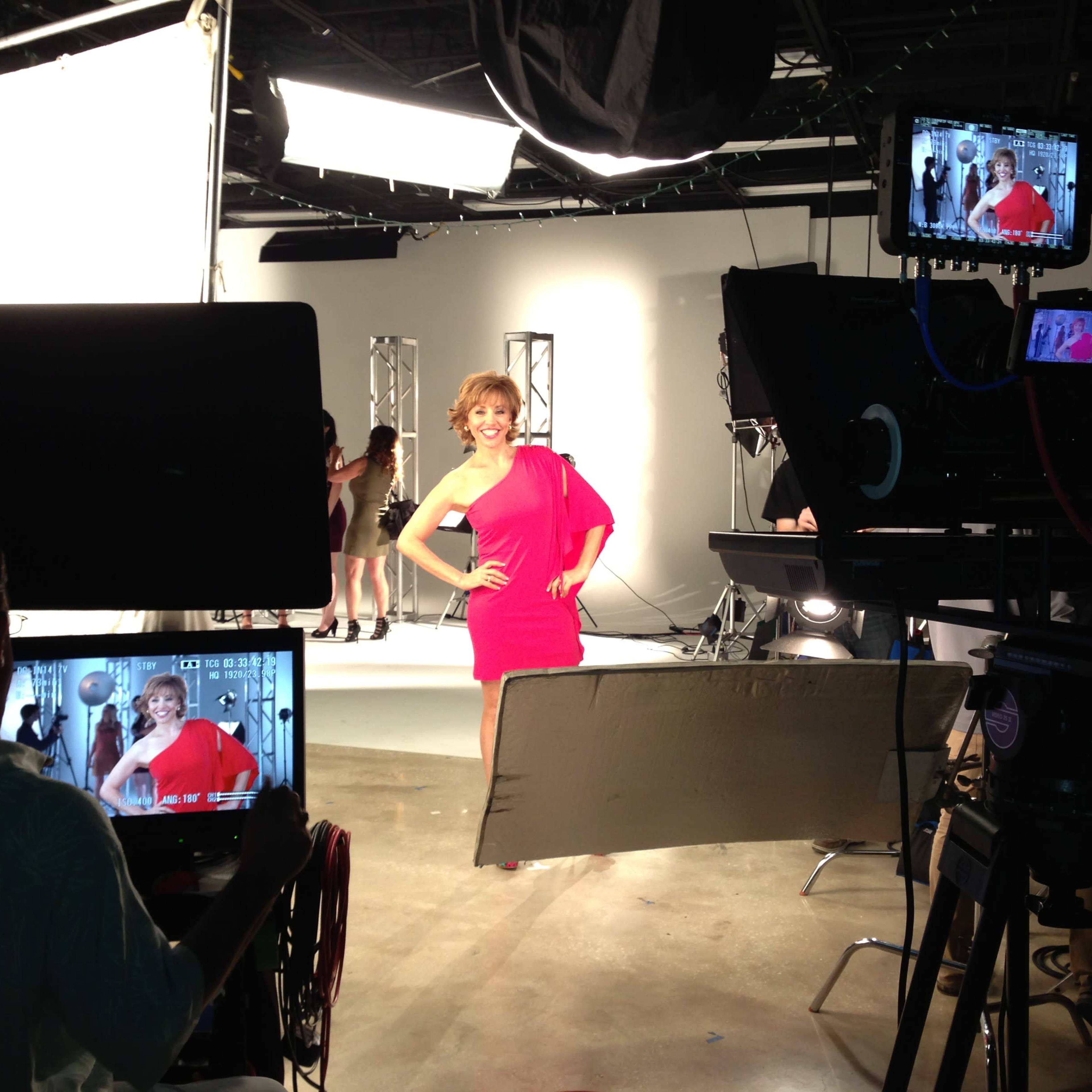 A woman in a bright pink dress poses confidently in a studio surrounded by cameras and lighting equipment.