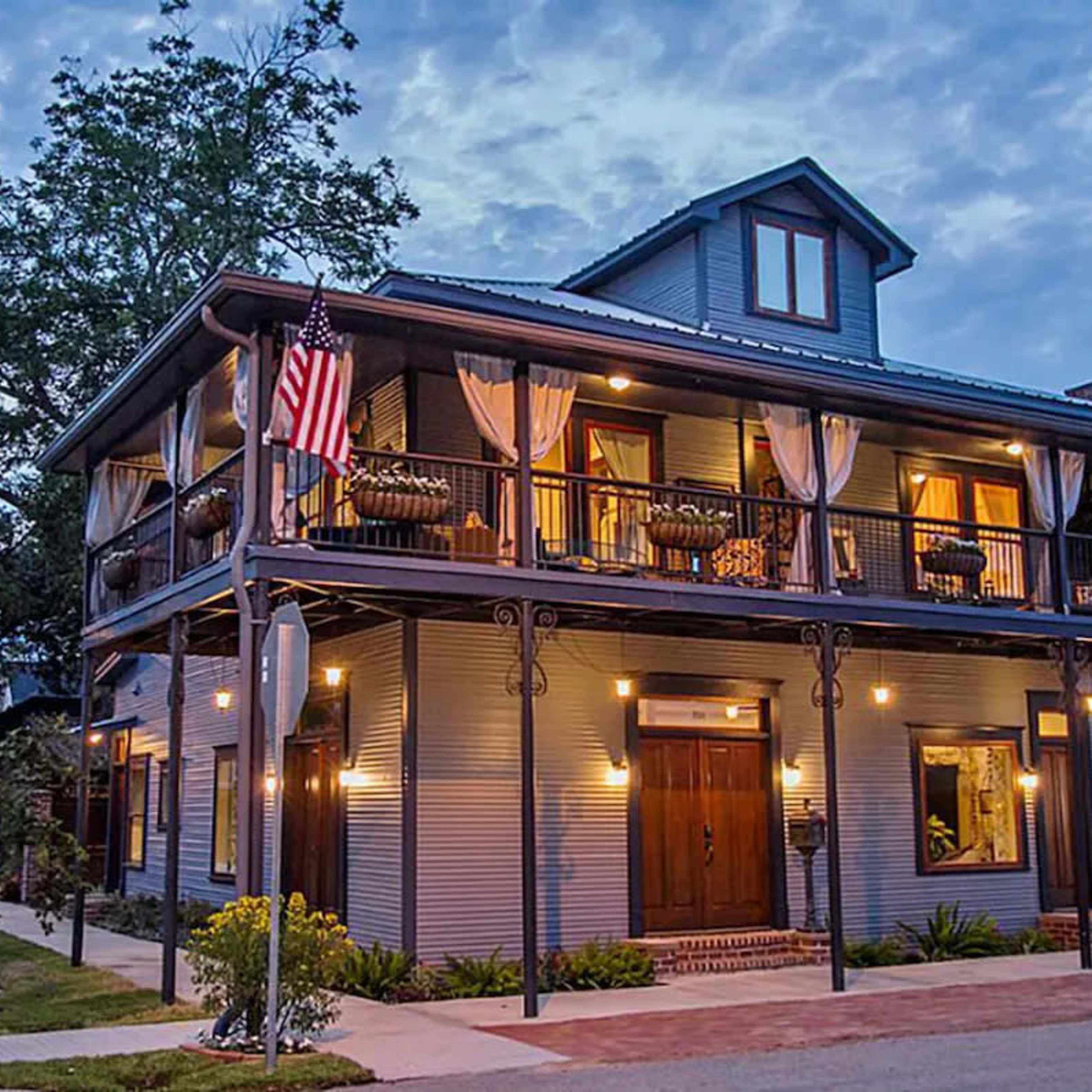 A large two-story gray house features multiple balconies, an American flag, and warm exterior lighting along a quiet street.