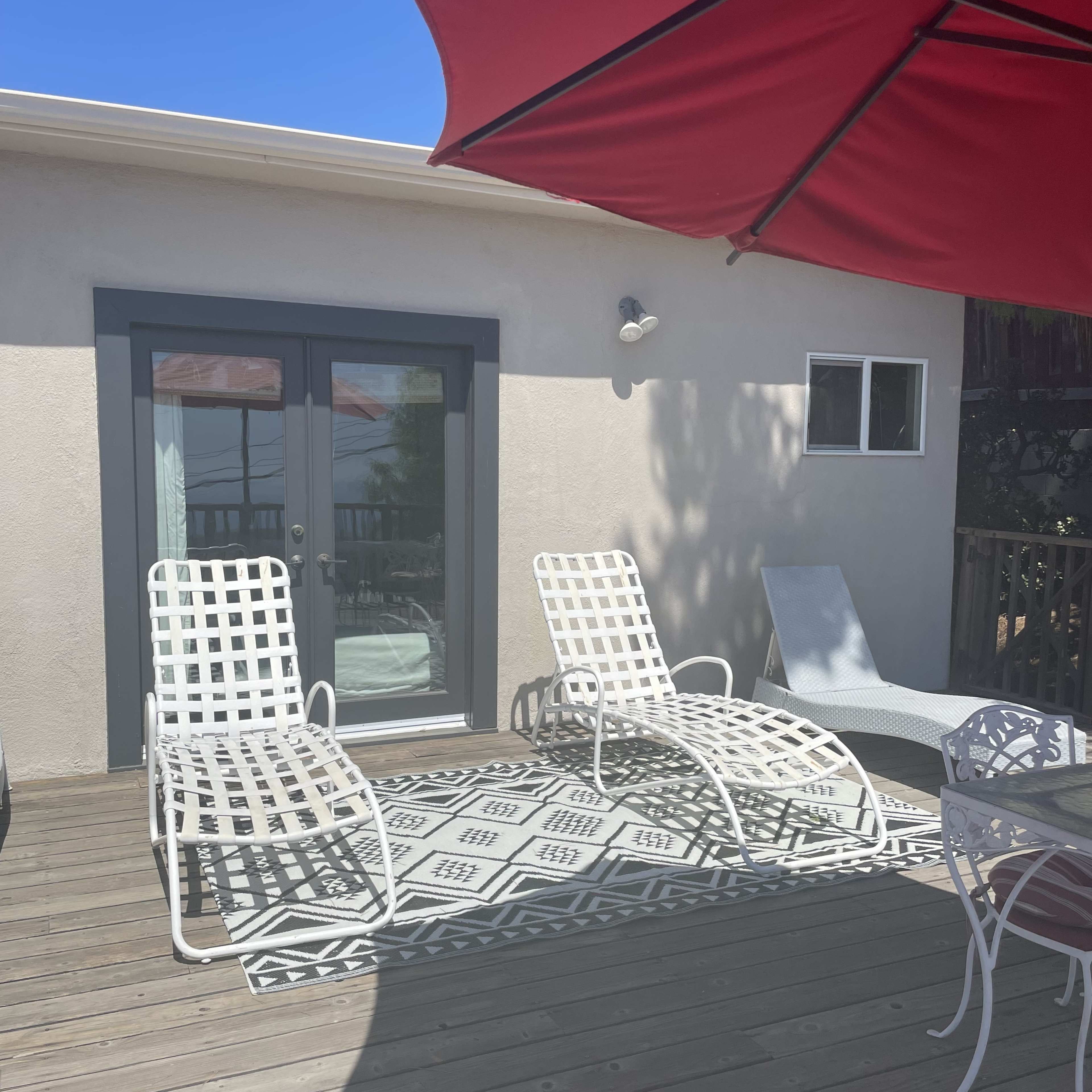 A sunny patio area with two white loungers, a small table, and a large red umbrella providing shade.