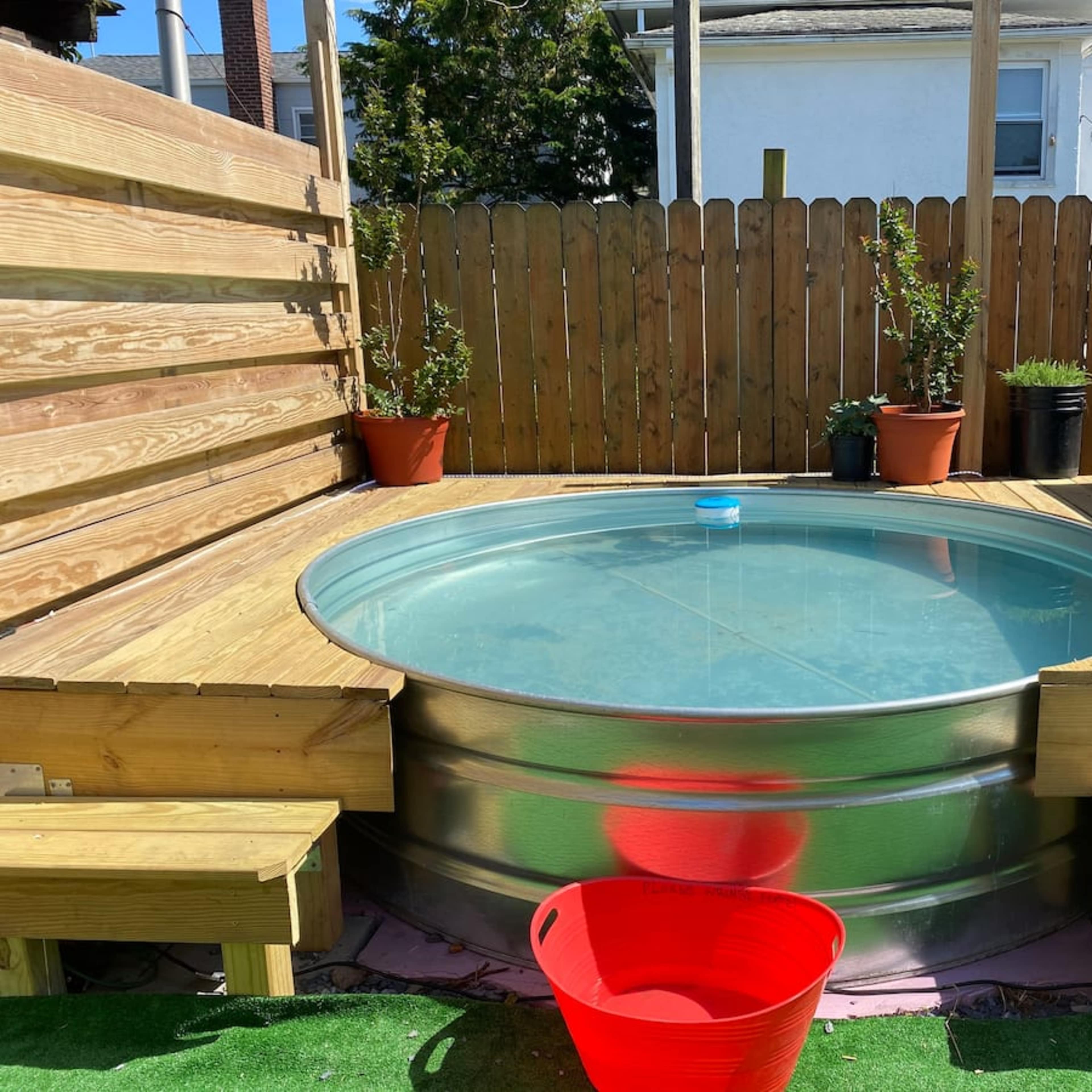 A circular, above-ground hot tub made of metal, surrounded by wooden benches and planters in a fenced outdoor area.