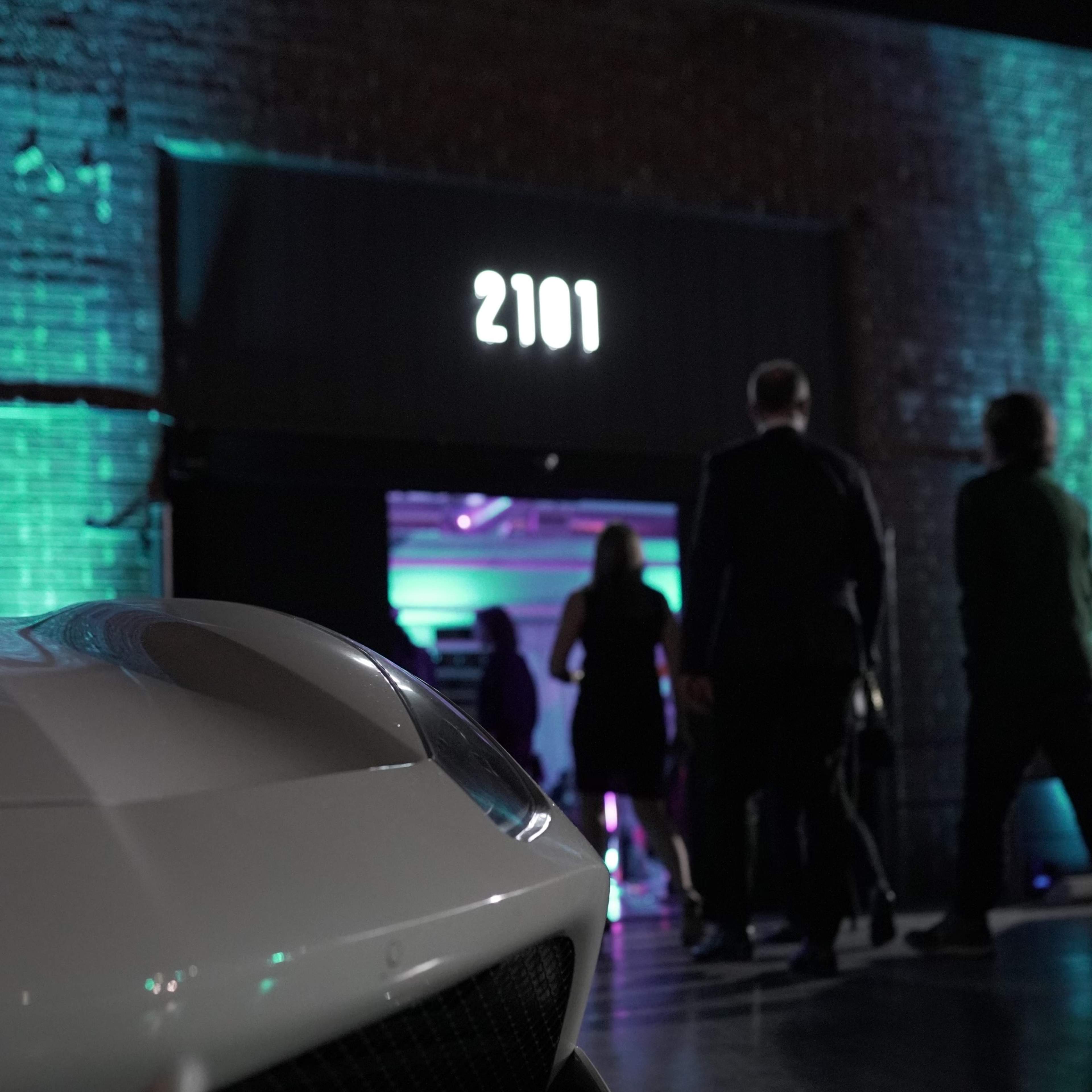 A sleek white car is parked in the foreground while people gather near a brightly lit entrance marked "2101."