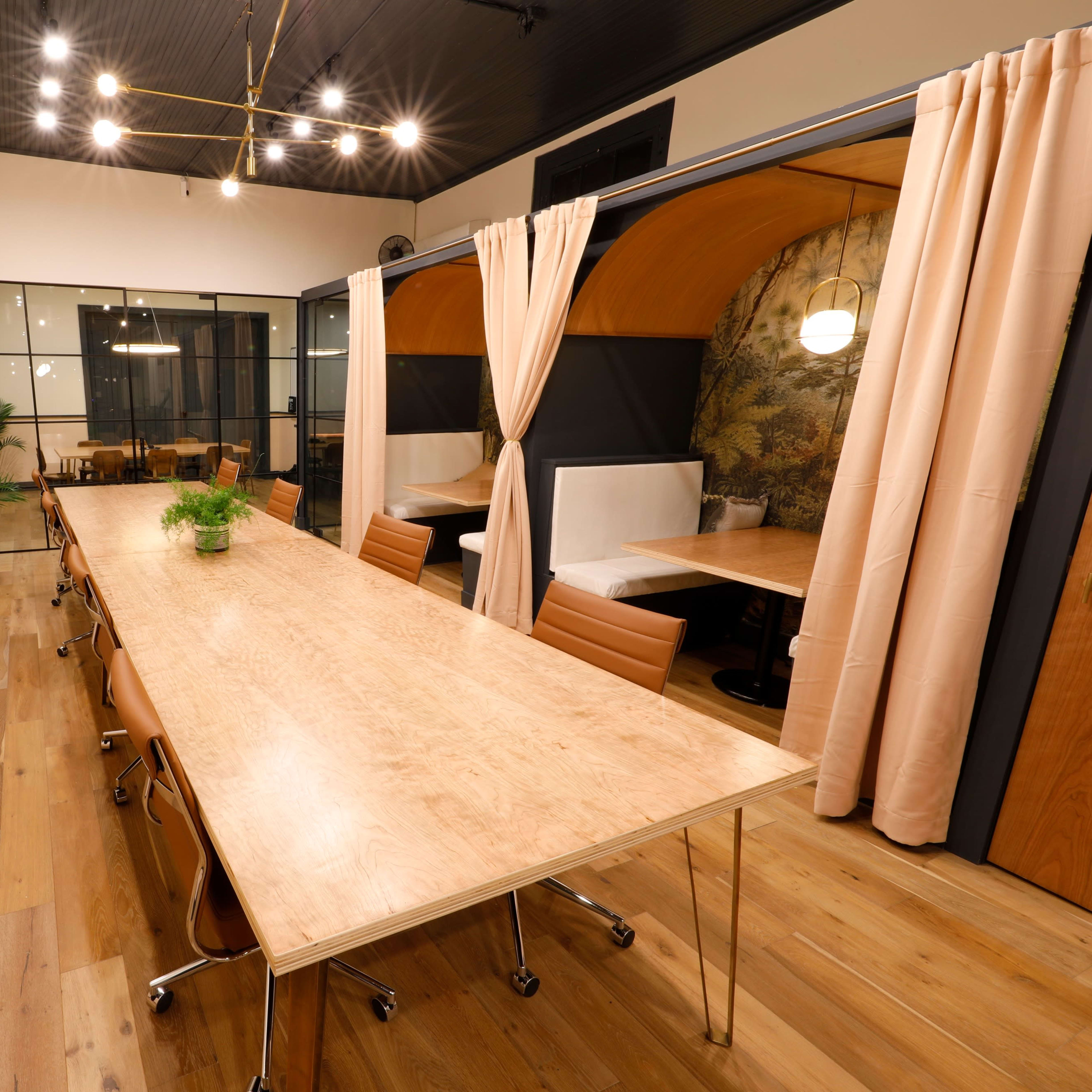 A large wooden table with rolling chairs is positioned in a well-lit meeting room featuring soft curtains and booths in the background.