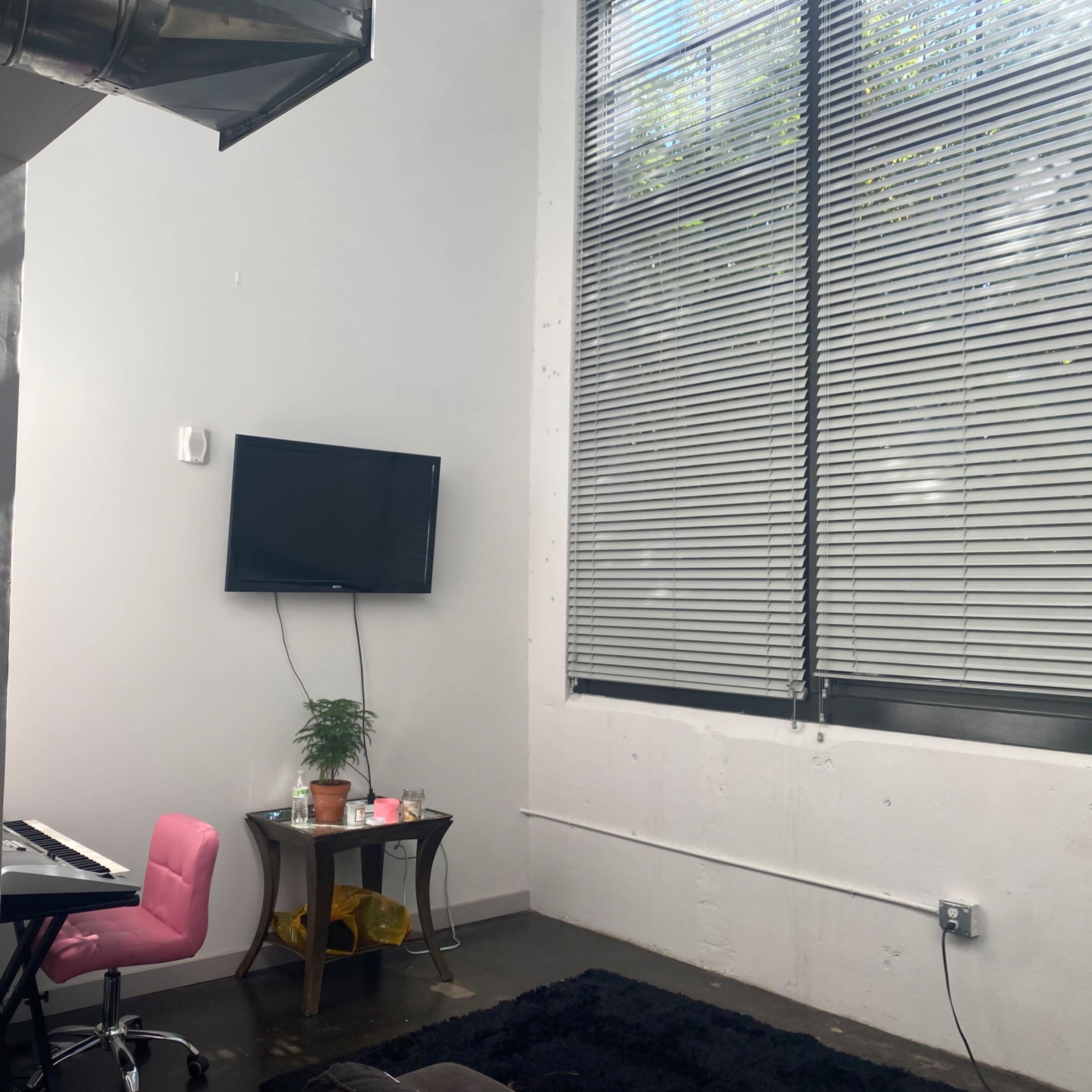 A small room with a keyboard near a pink chair, a plant on a table, and a wall-mounted television beneath large windows with blinds.