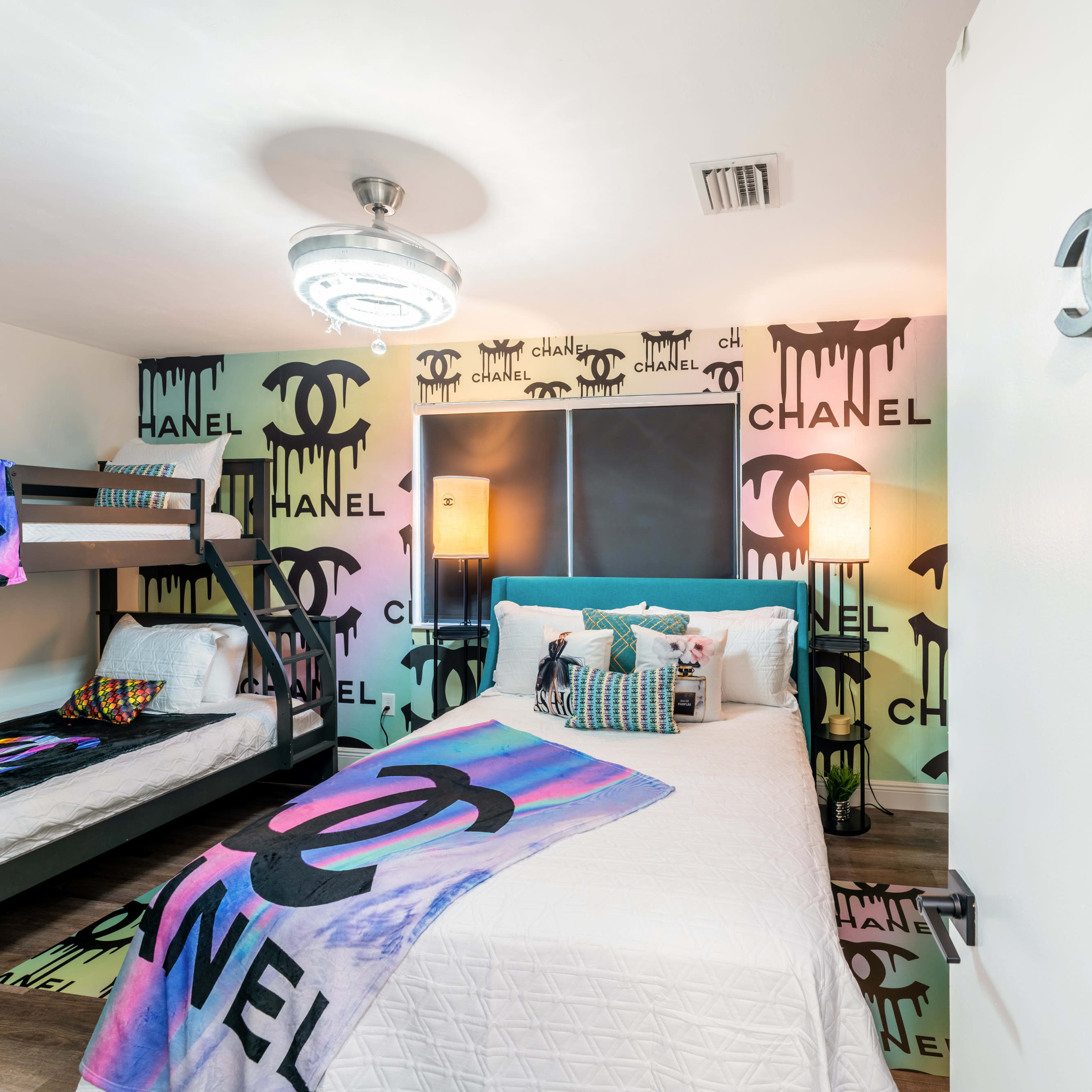 A brightly decorated bedroom featuring a bunk bed, a single bed, and walls adorned with colorful Chanel logos.