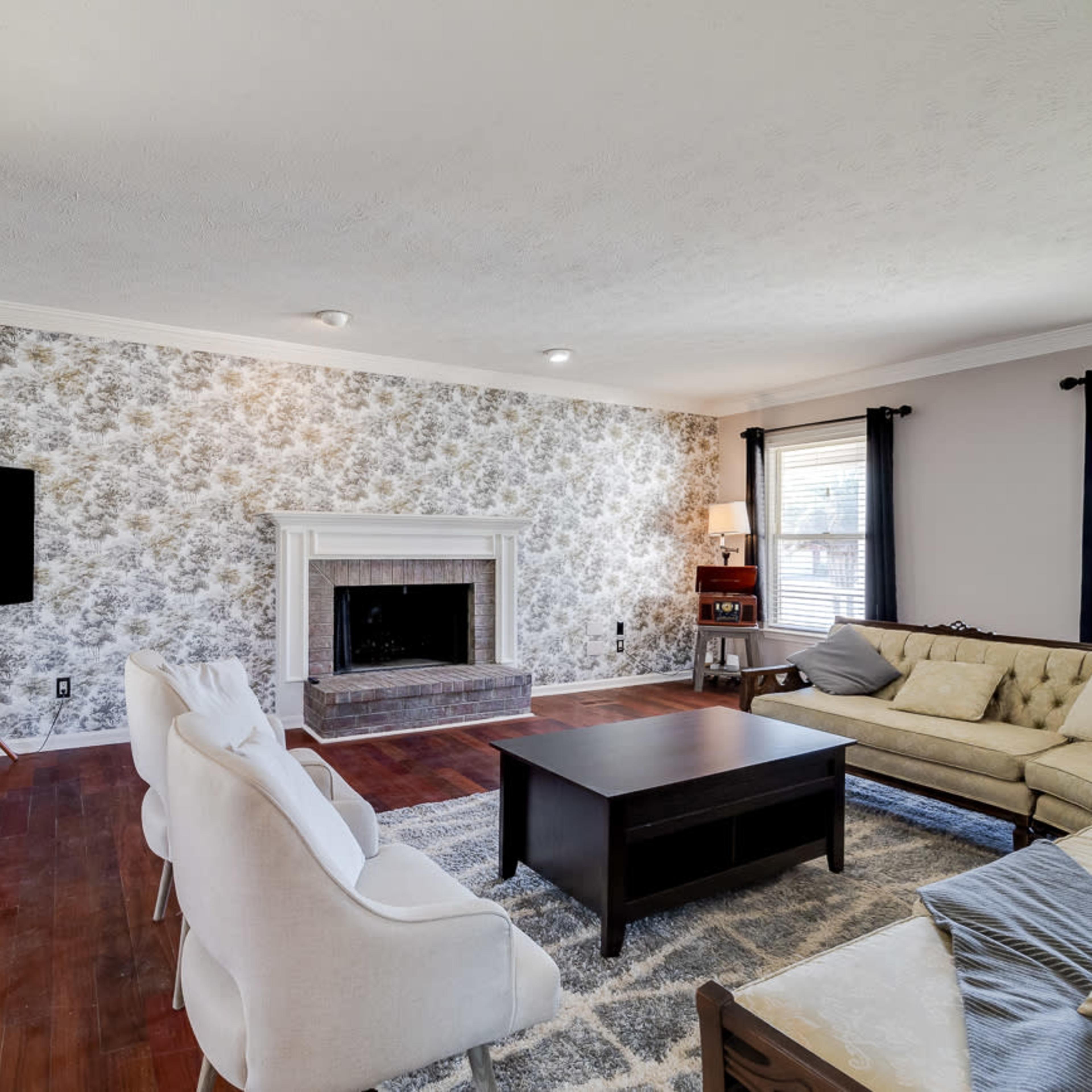 The living room features a fireplace against floral wallpaper, a flat-screen TV on a stand, a beige sectional sofa, and a coffee table on a rug.