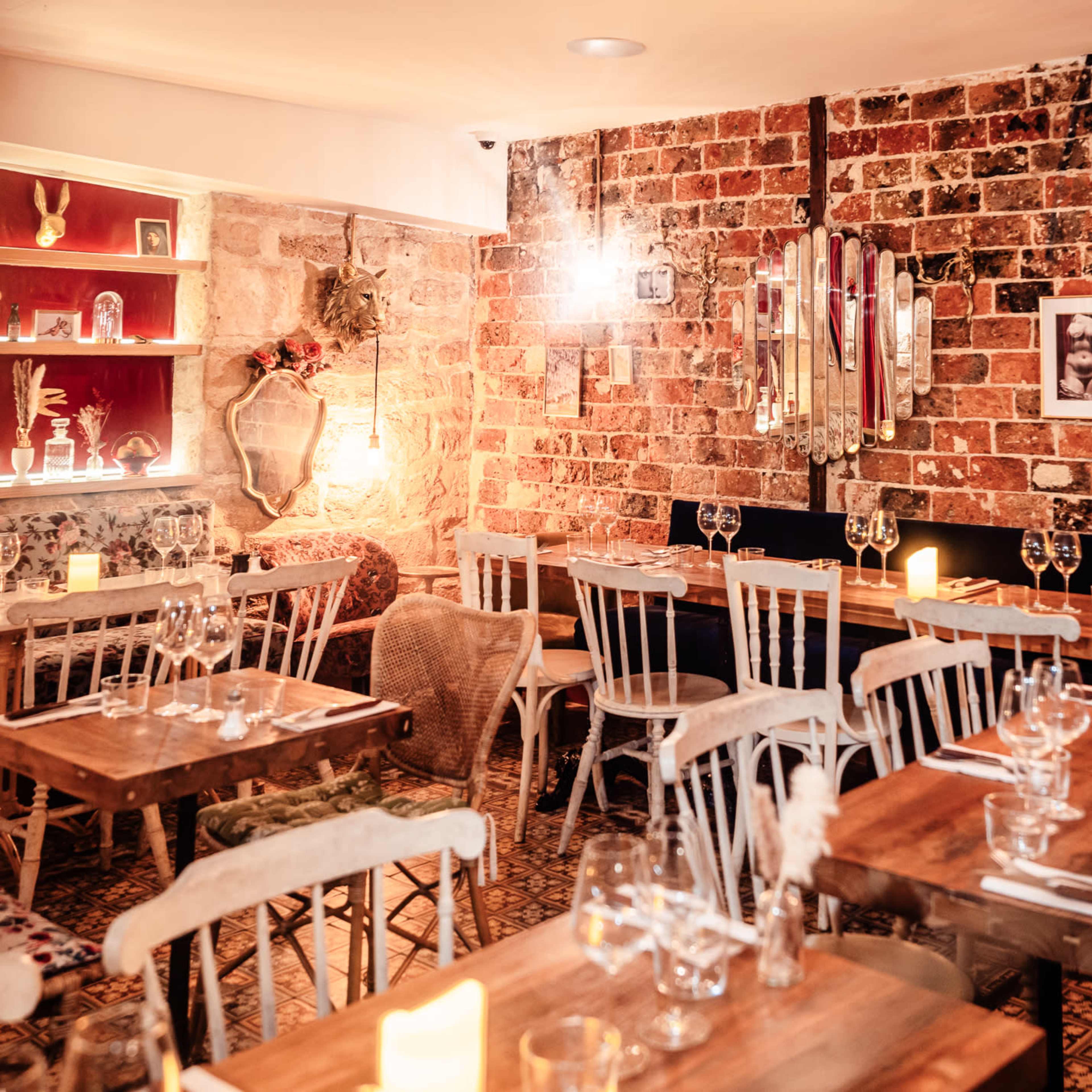 An intimate restaurant featuring wooden tables set with glassware, a brick wall adorned with mirrors, and warm lighting creating a cozy atmosphere.