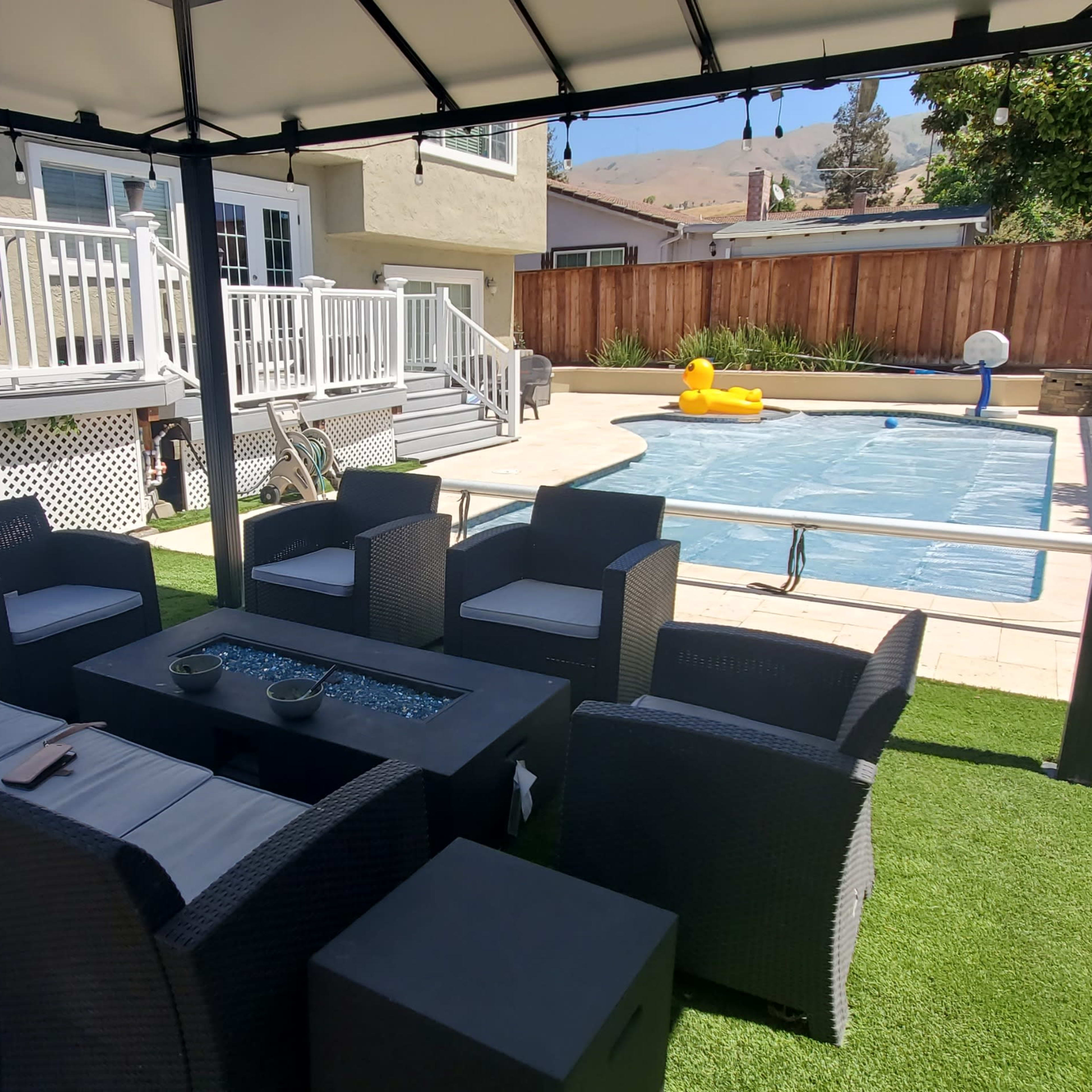 A shaded outdoor seating area with black wicker furniture surrounds a glass table, overlooking a pool with a yellow float.