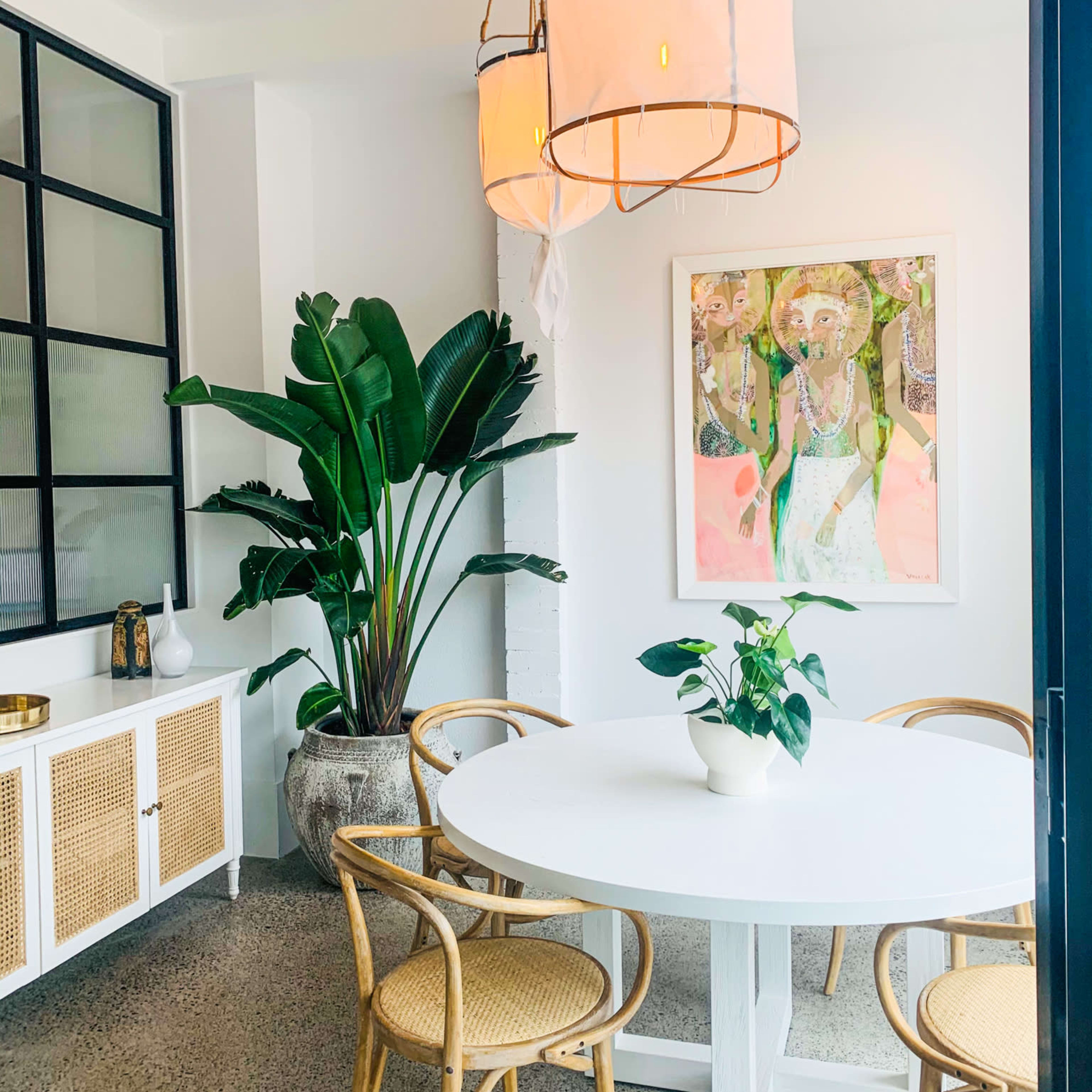 A dining area features a round white table surrounded by four rattan chairs, with large plants and artwork on the walls.