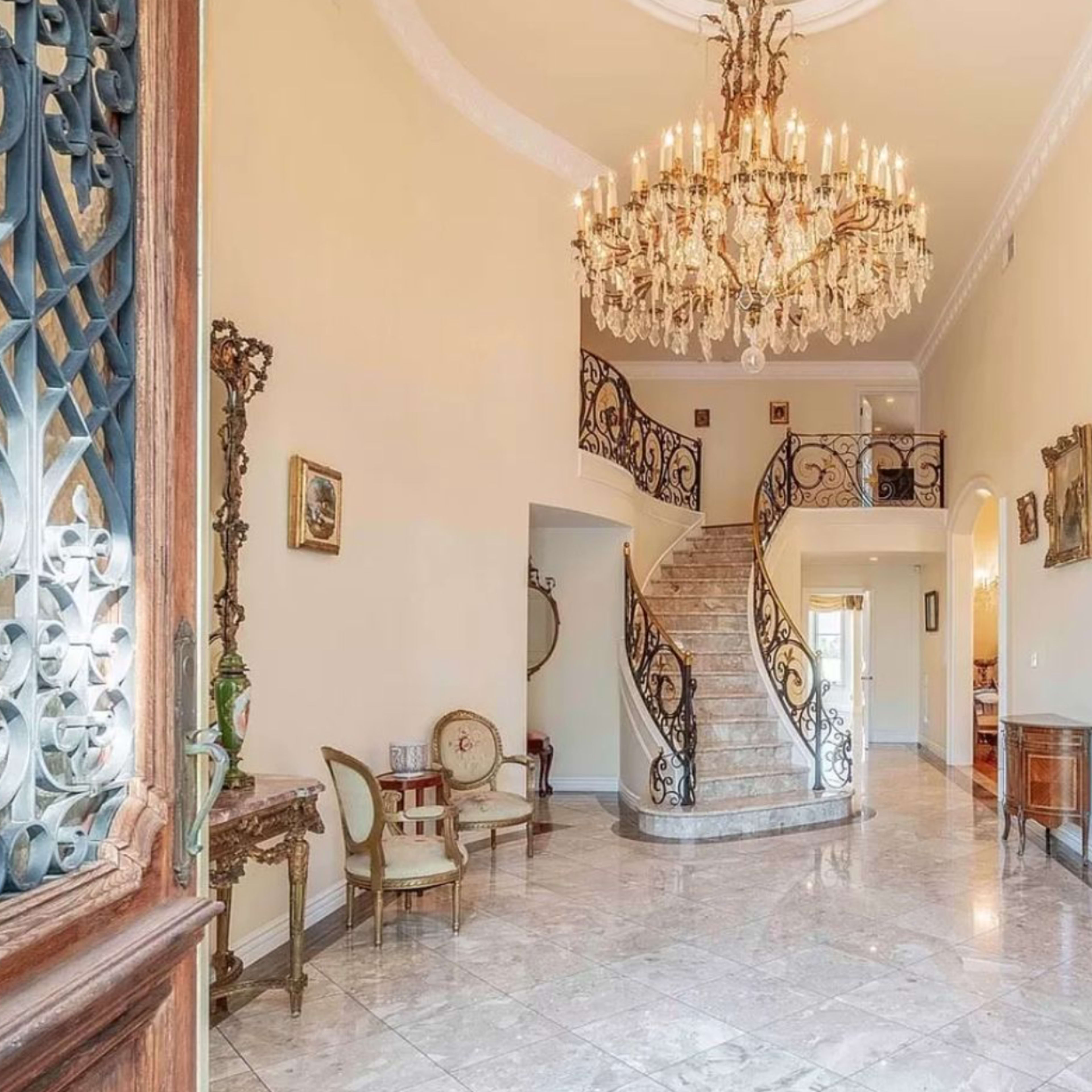 An elegant foyer featuring a grand chandelier, a winding staircase with ornate iron railings, and marble flooring.
