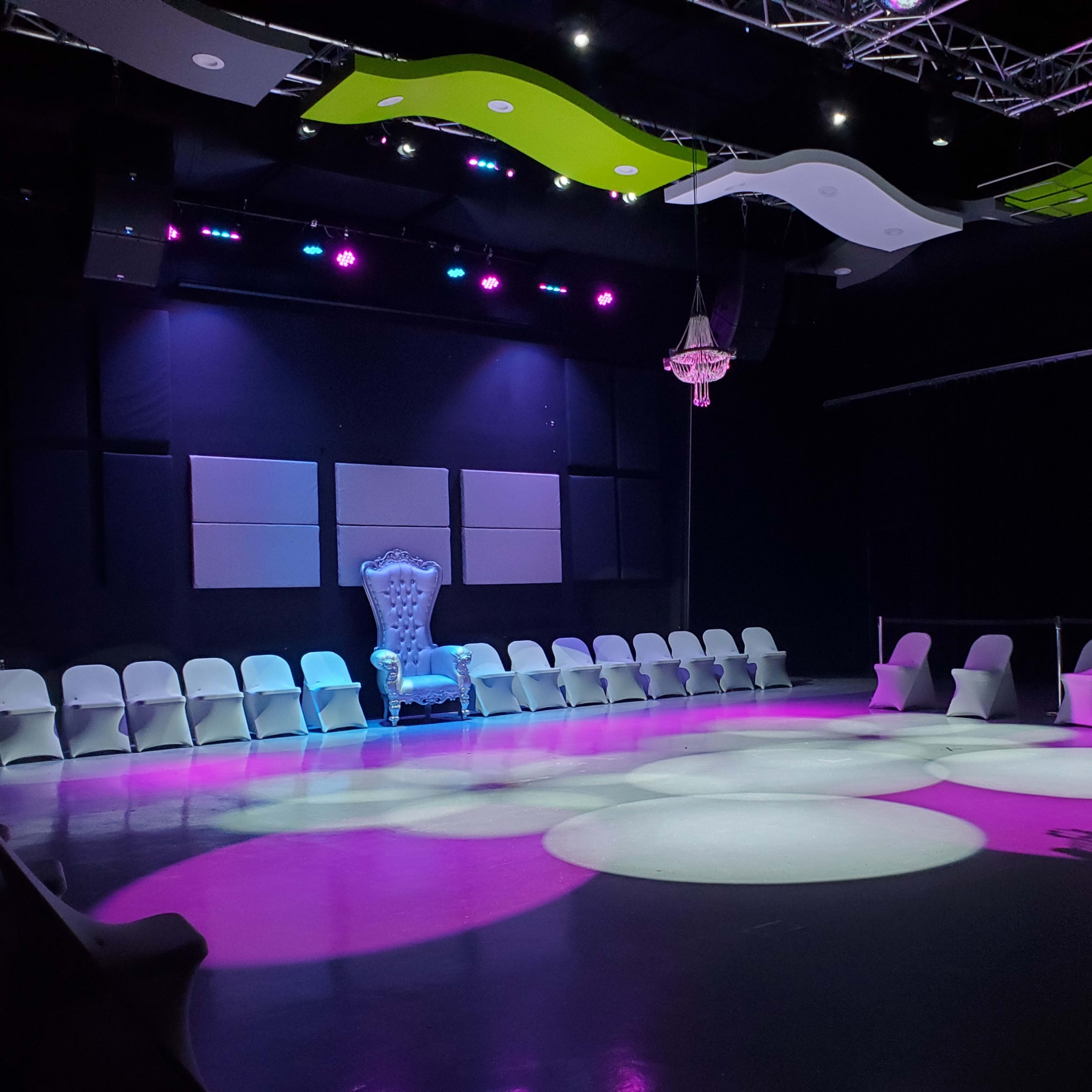 A dimly lit event space with arranged white chairs surrounding a circular area, featuring a large blue throne under purple and green lighting.