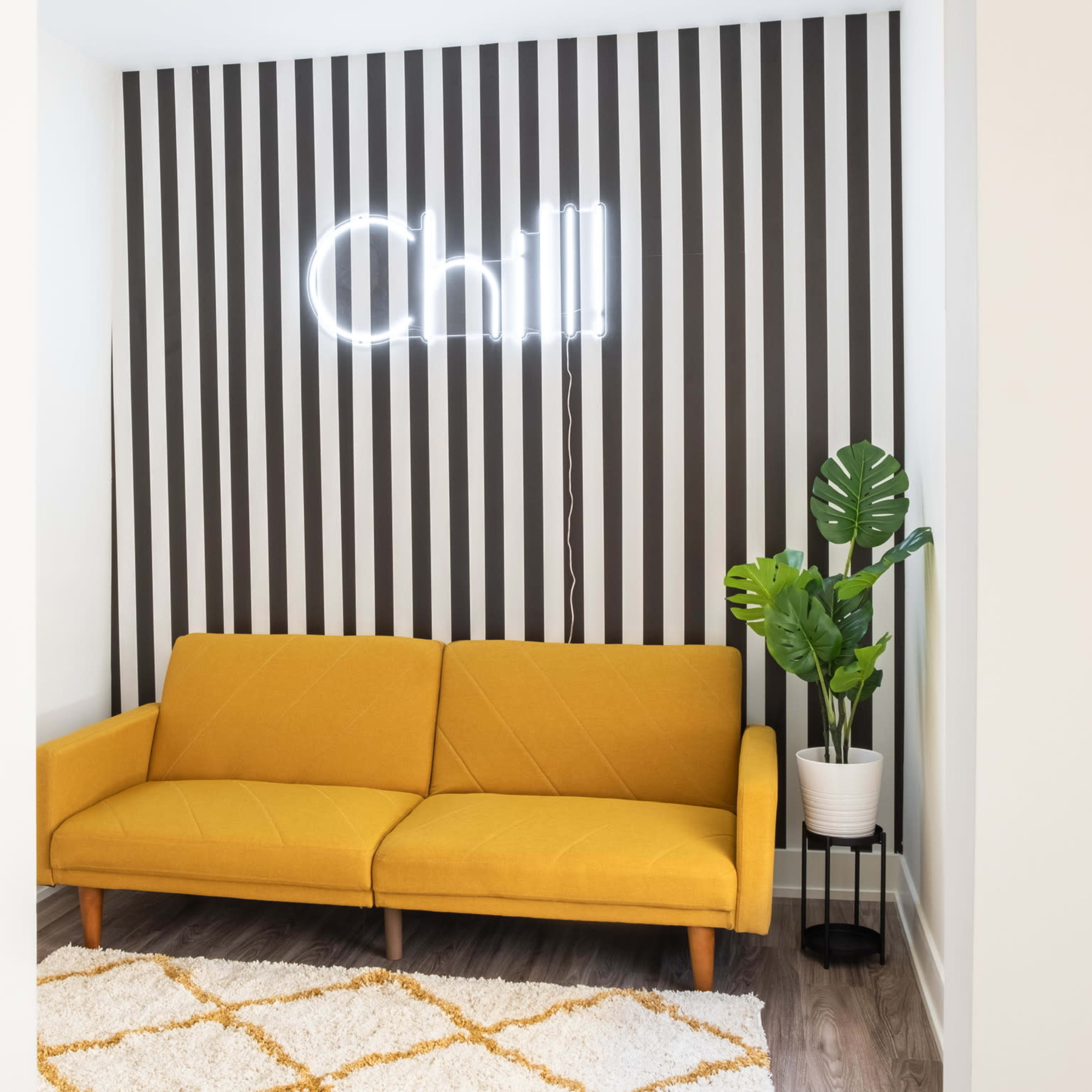 A yellow sofa sits in a room with striped walls, a neon sign that reads "Chill," and a potted plant in the corner.