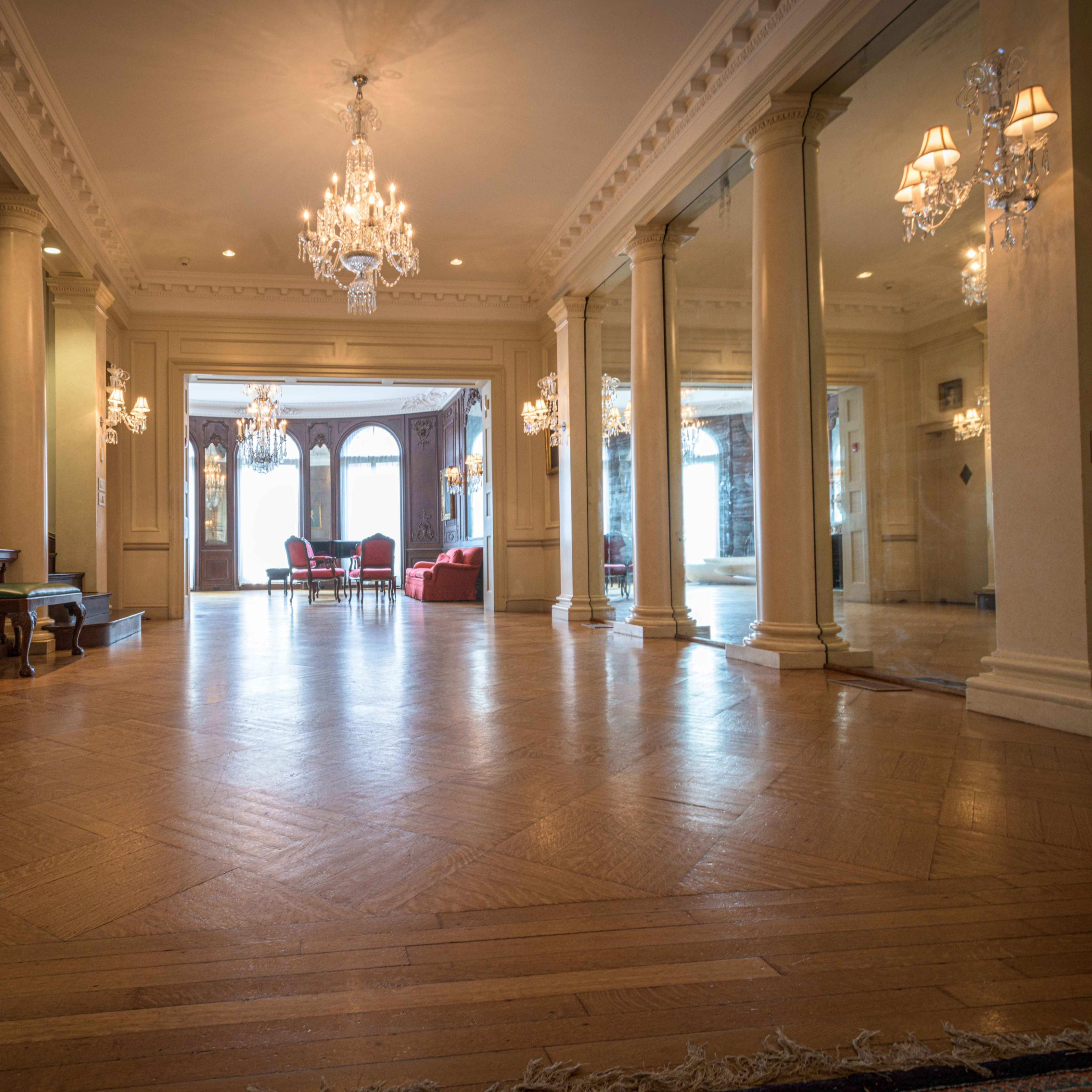 A spacious foyer with polished wooden floors, tall columns, and elegant chandeliers, leading to a living area with red furniture.