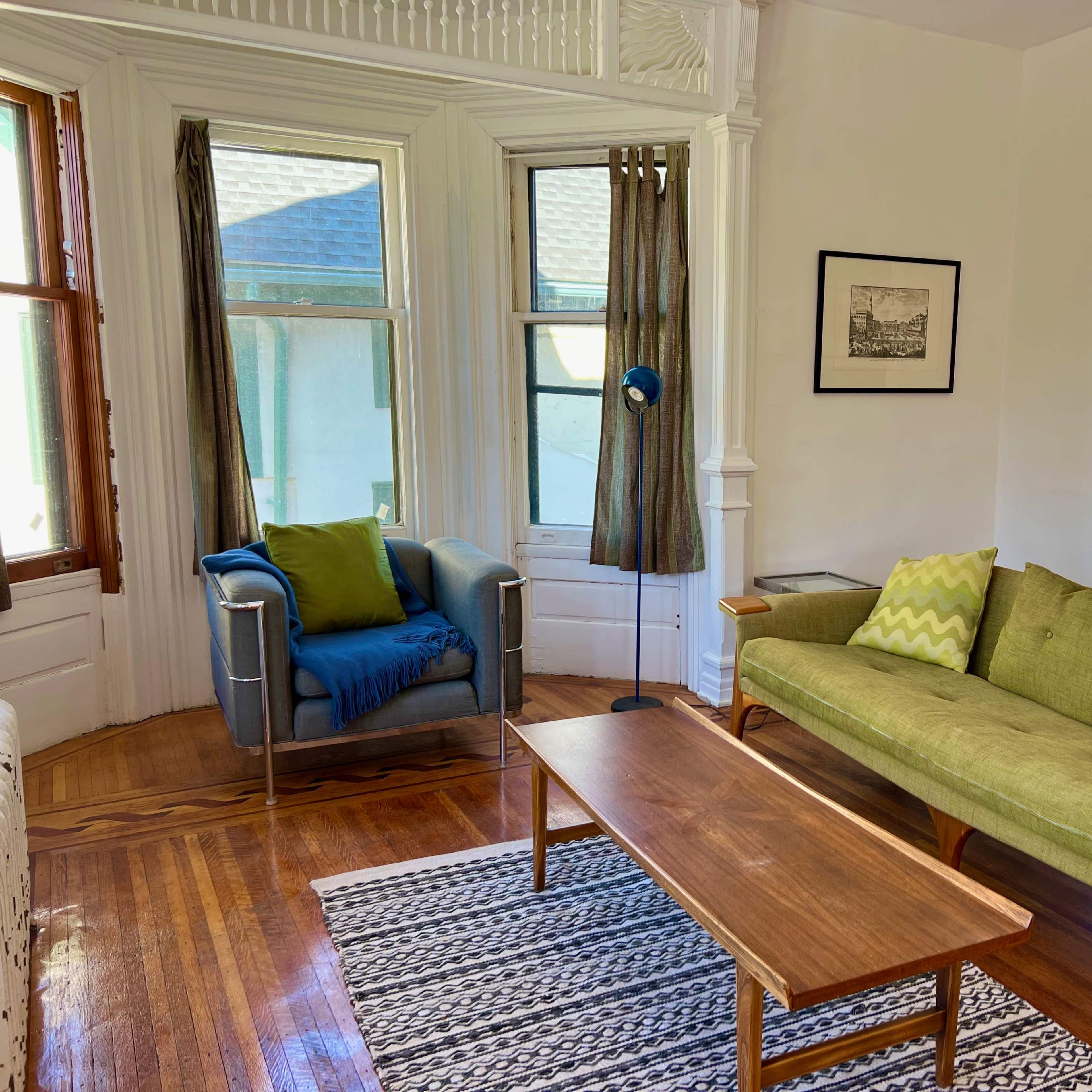 A bright living room with a green sofa, a blue armchair, a wooden coffee table, and two large windows with curtains.
