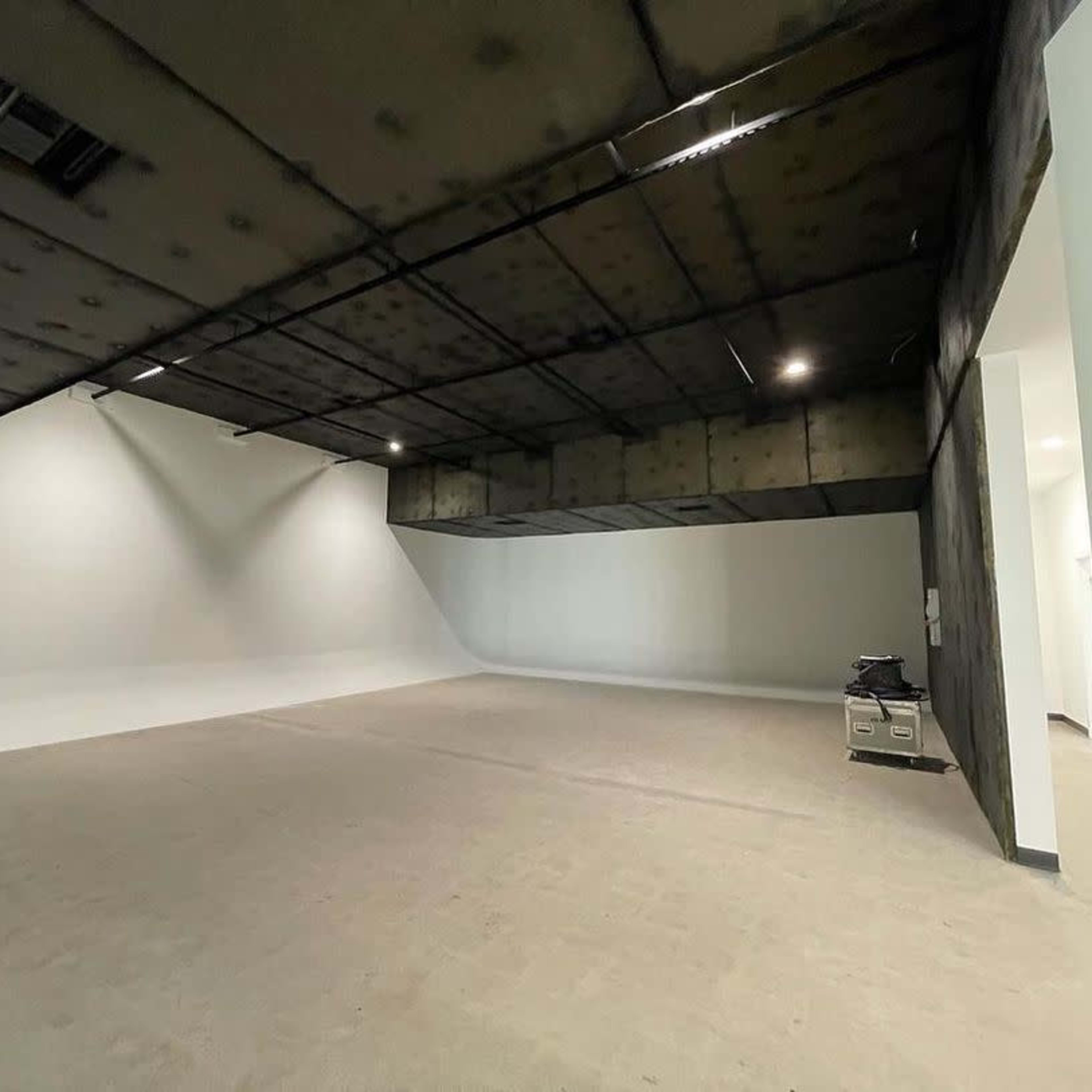A spacious, empty room with a smooth white wall and a ceiling that is partially blackened, illuminated by overhead lights.