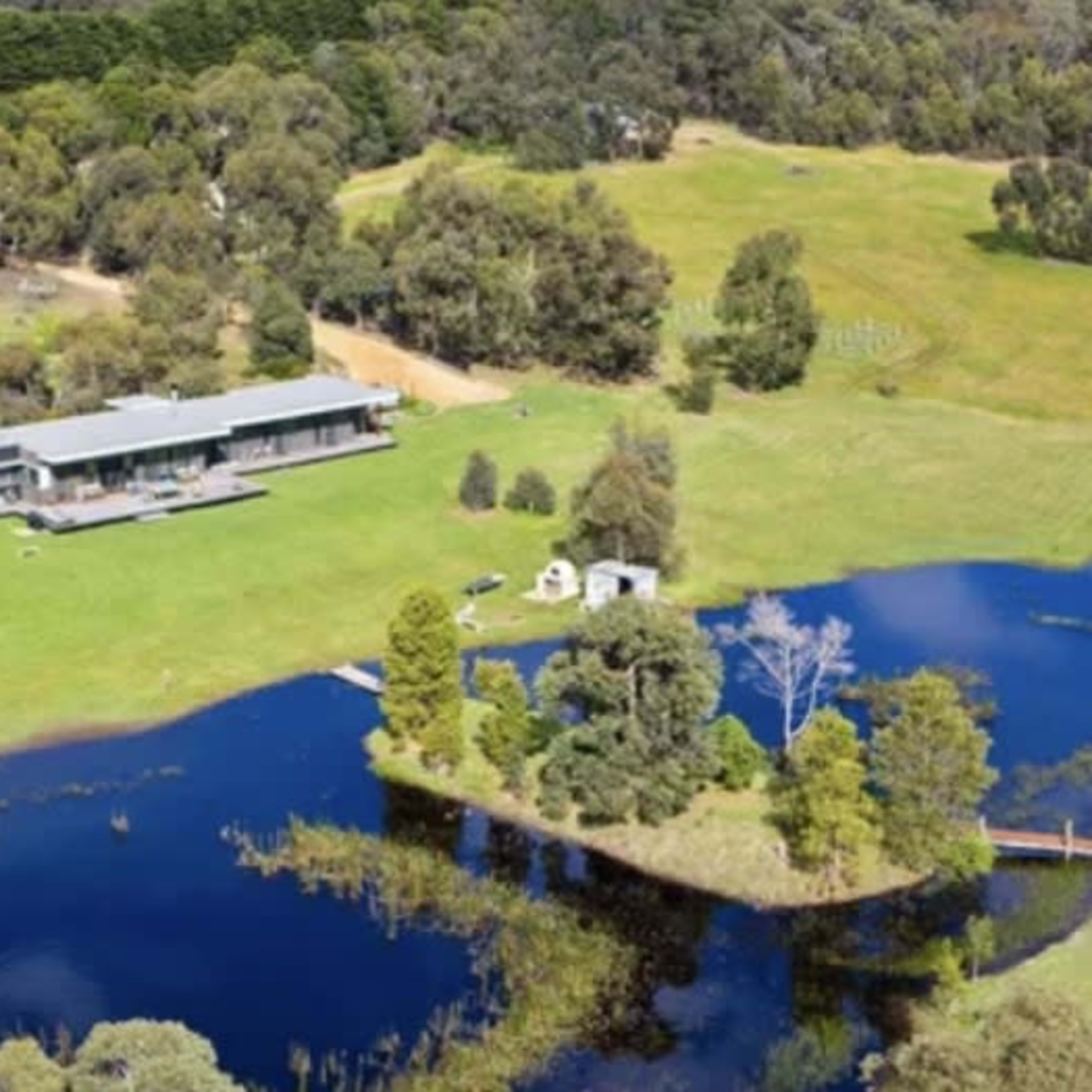 A modern house situated next to a pond in a green, open landscape surrounded by trees.
