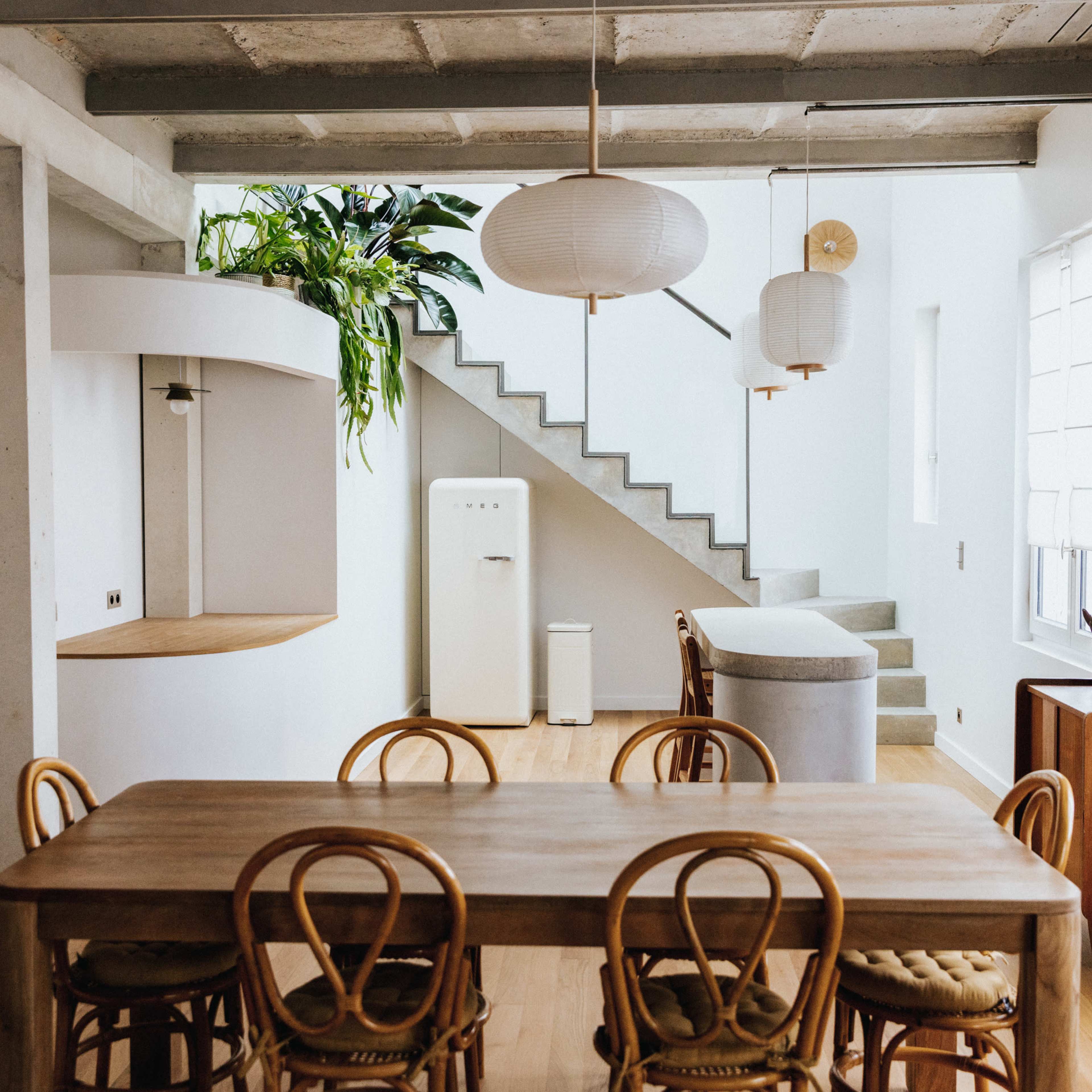 A modern dining area features a wooden table surrounded by chairs, with a staircase in the background leading to an upper level, illuminated by hanging pendant lights.