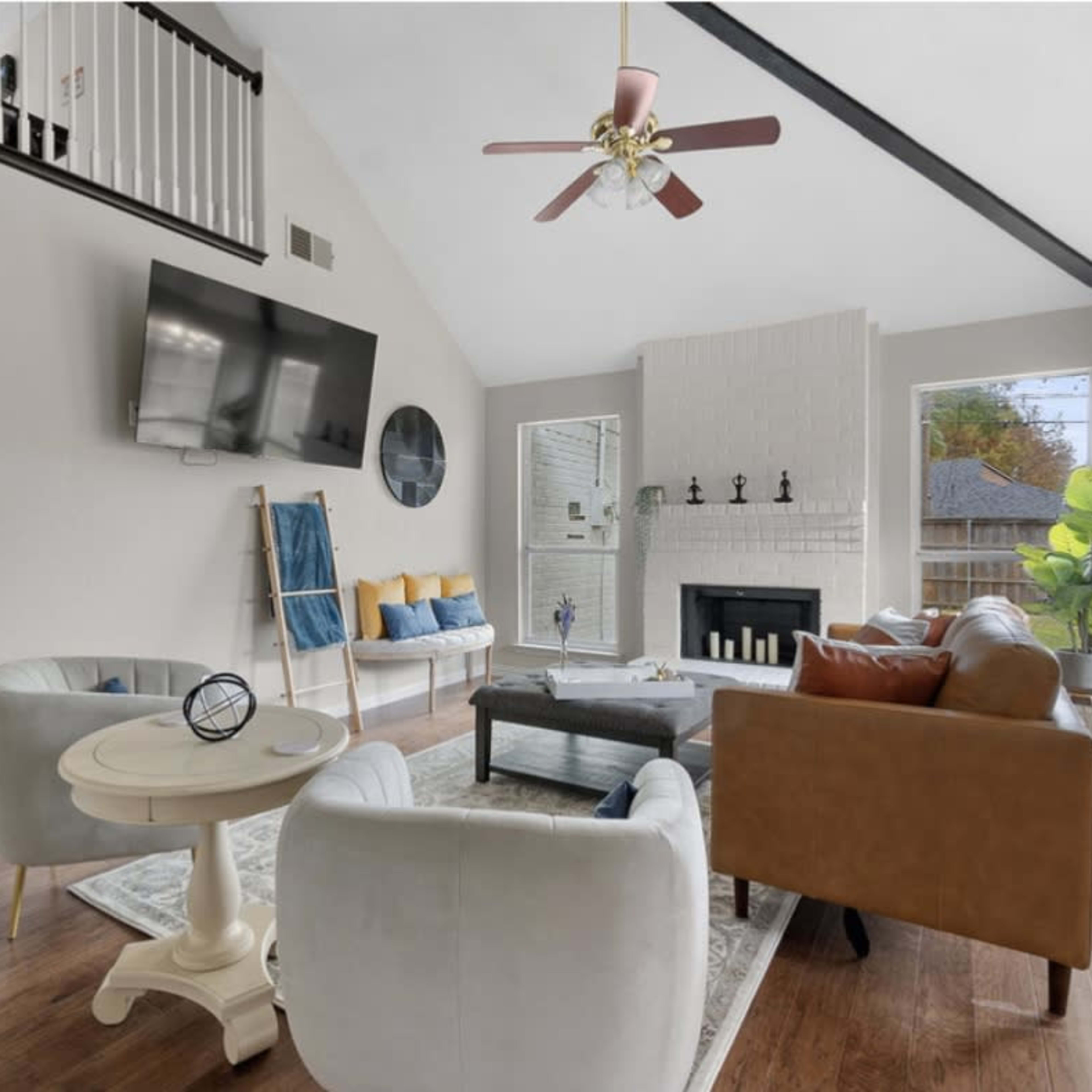 A bright living room features a large window, a flat-screen TV on the wall, and a mix of modern furniture including a coffee table and two sofas.