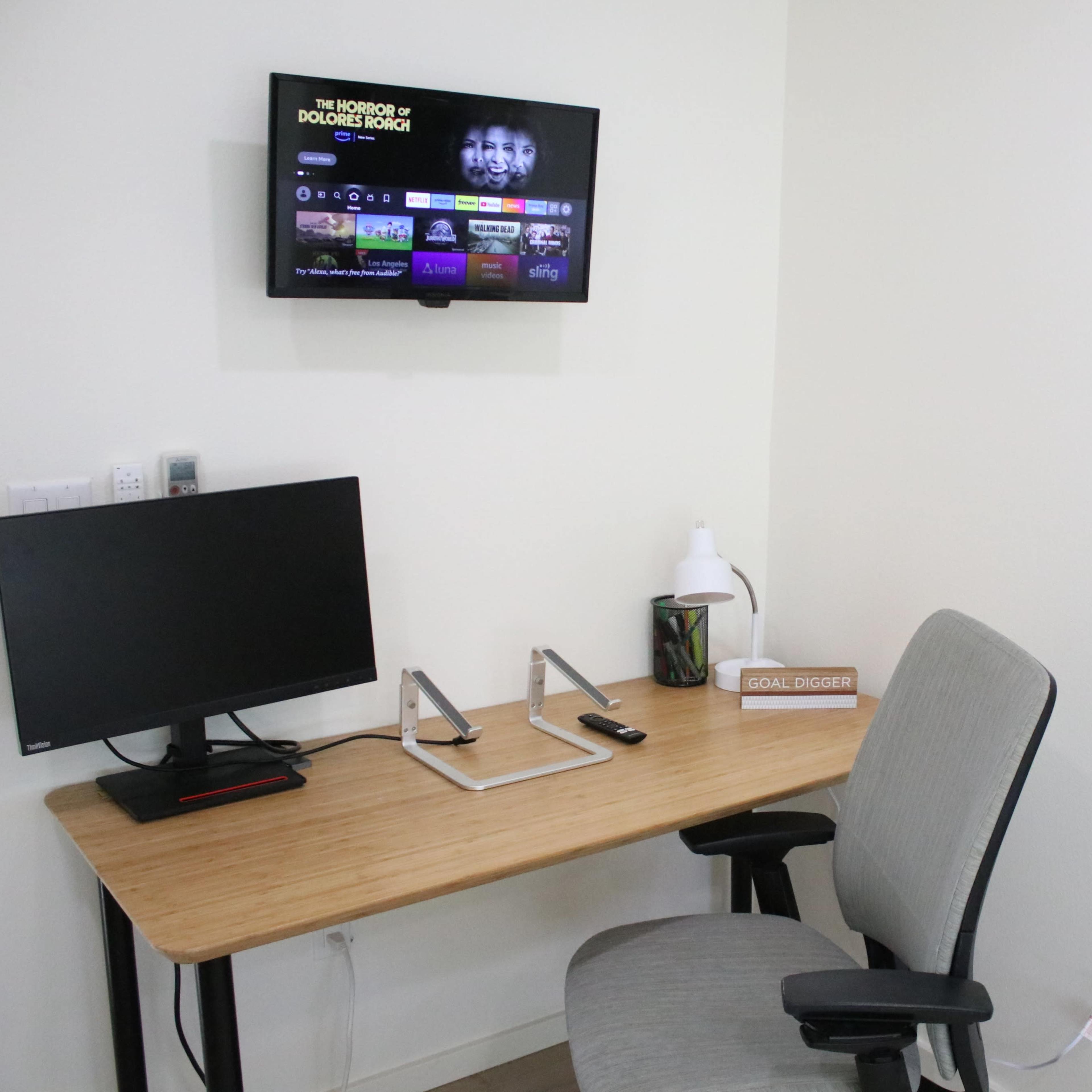 A wooden desk with a computer monitor and a chair is positioned under a wall-mounted television in a minimalistic room.