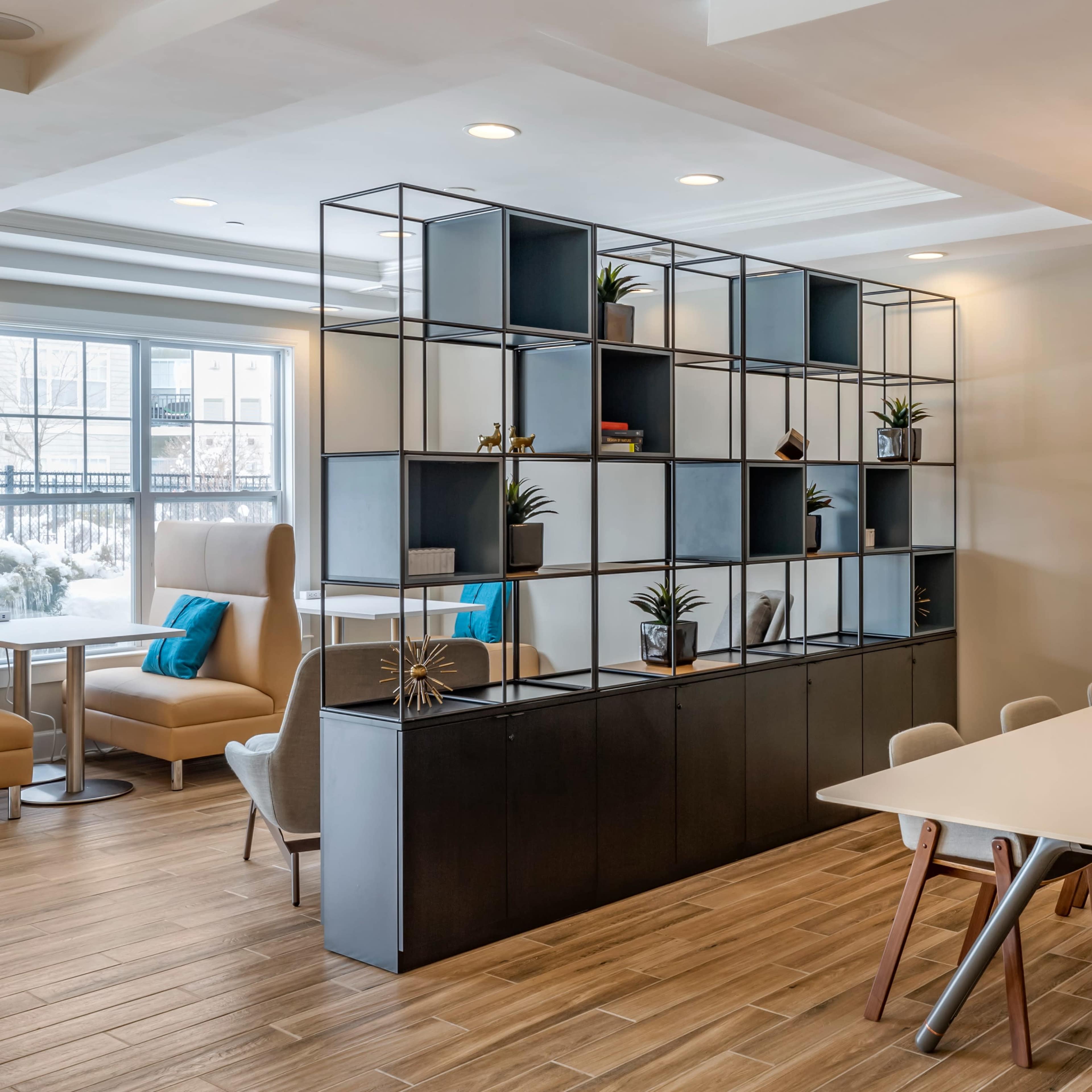A modern dining area with a long table, light-colored chairs, and a geometric shelving unit filled with plants and decorative items.