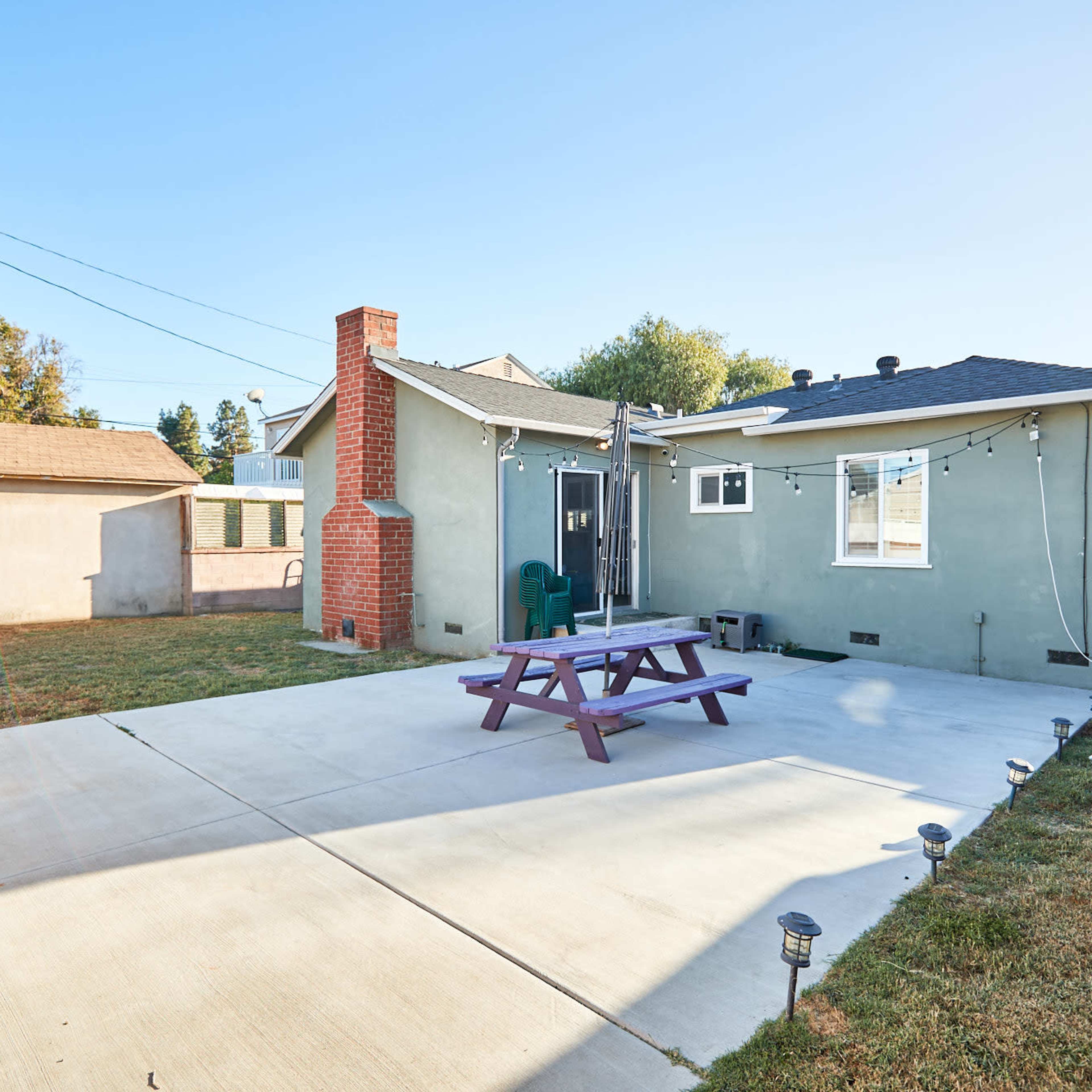 A backyard with a concrete patio, a purple picnic table, a brick chimney, and several trees and grass areas in the background.